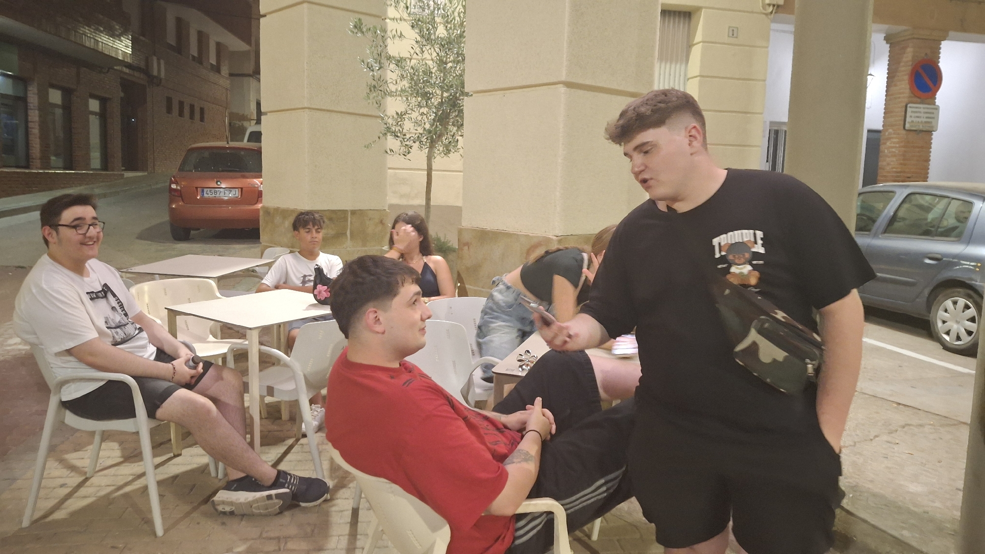 The image depicts an outdoor seating area at a cafe or restaurant in Híjar, Spain. In the foreground, a young man wearing a black t-shirt with a graphic print and a camouflage crossbody bag is engaged in conversation with another young man seated in a white chair, who is wearing a red t-shirt. Both men have dark hair and appear to be in their late teens or early twenties.
Behind them, other individuals are visible. To the left, a young man with glasses and a white t-shirt is seated at a table, looking to his left and smiling. Further back, another young man in a white t-shirt is seated. A young woman with long dark hair is seated in the background, covering her face with her hand. Another young woman in a dark top and denim shorts is leaning over a table, holding a mobile phone.
The environment includes several white plastic chairs and tables, typical of outdoor cafes. In the background, a light-colored building with architectural columns and some greenery, including a small tree, are visible. The street is paved, and an orange car with a Spanish license plate is parked on the left side of the image. A light blue car is parked on the right side, near a no parking sign. The lighting suggests it is evening or nighttime.