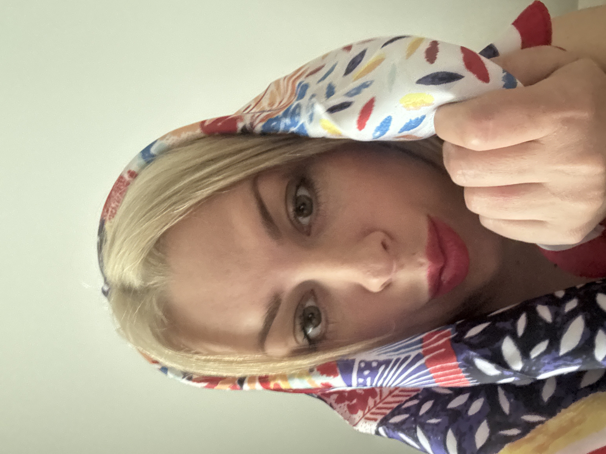 A woman with blonde hair and red lipstick is looking at the camera and holding a colorful scarf to her head. The background is a plain, light-colored wall. There are no discernible landmarks or environmental features that would indicate the location is Sochi, Russia.