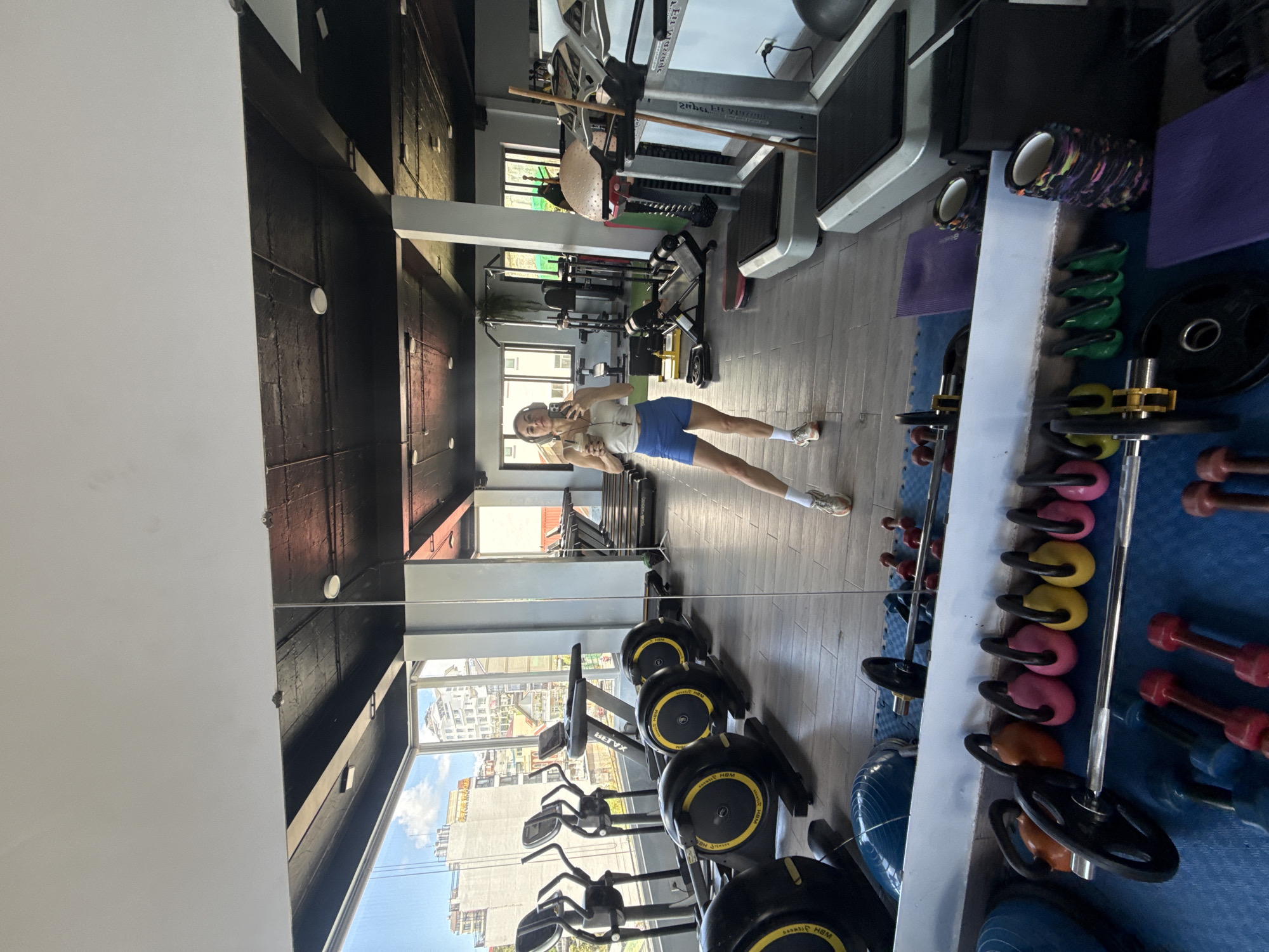 A woman is taking a selfie in a gym located in Da Lat, Vietnam. She is standing in front of a large mirror, wearing a white tank top, blue shorts, white socks, and athletic shoes. She is holding a phone in her right hand and a coffee drink in her left hand, and she has headphones on. The gym is well-equipped with various exercise machines, including treadmills, elliptical trainers, and weight machines. Dumbbells, kettlebells, and weight plates are neatly arranged on the floor and on racks. The gym has a modern design with wooden floors and a black ceiling. Large windows offer a view of the city, with buildings and trees visible in the background. The overall atmosphere appears to be clean and well-maintained.