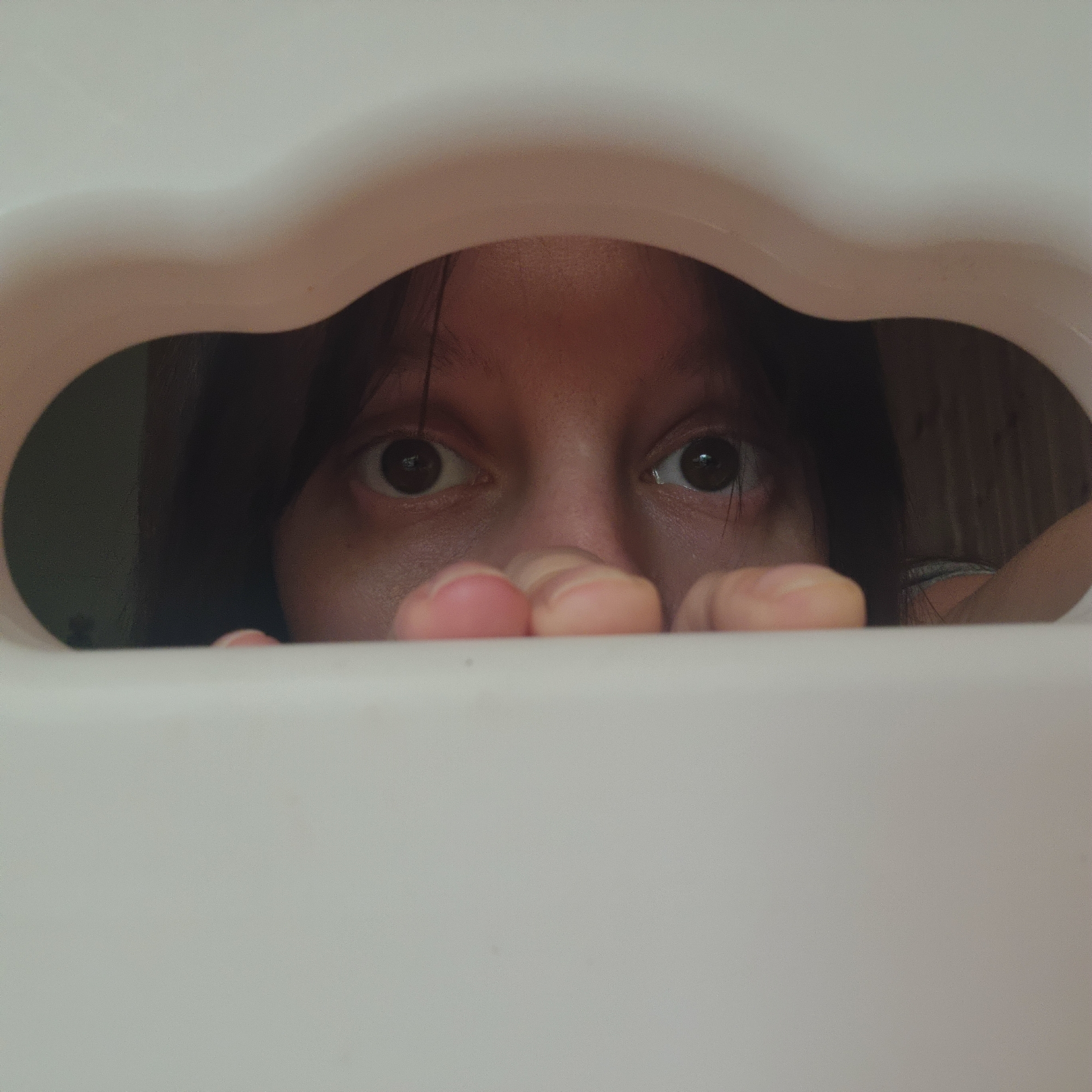 A close-up shot shows a child peeking through a white, cloud-shaped opening. The child's eyes and forehead are visible, with dark hair framing their face. Their hands are resting on the edge of the opening, with their fingers curled. The background is out of focus, but appears to be indoors with wooden paneling on the right side. The image is taken from a low angle, looking up at the child. The lighting is soft and diffused.