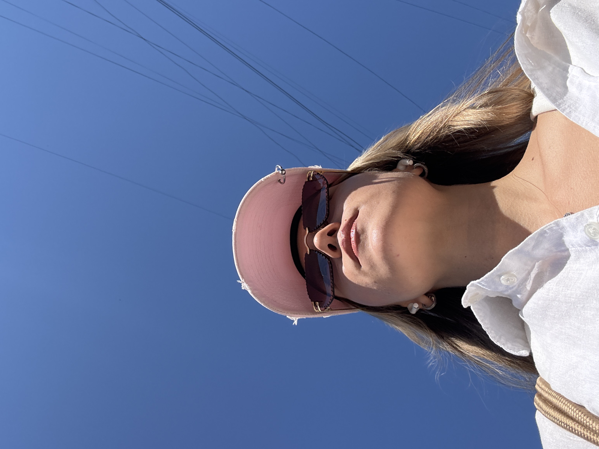 A low-angle, medium shot captures a young woman from the chest up, looking upwards against a clear, vibrant blue sky. She is wearing a light pink baseball cap with frayed edges, tilted forward to partially obscure her forehead. Dark, angular sunglasses with gold accents rest on her nose. Her lips are slightly parted and appear to have a subtle gloss. She has blonde hair that falls past her shoulders and is wearing a white collared shirt, partially unbuttoned. Small, hoop earrings are visible in her ears, one with a white charm. A strap of a tan handbag is visible over her left shoulder. The sky is mostly clear, with a few thin, dark power lines crisscrossing the upper right portion of the frame. The lighting suggests a sunny day.