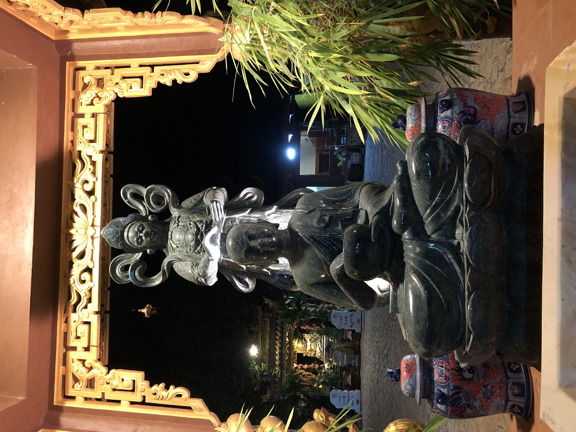 This is a photograph taken at night in Mũi Né, Vietnam. The main subjects are a large, dark green stone statue depicting a multi-headed figure with serene facial features, possibly a deity or enlightened being, and a large ornate golden gate or archway in the foreground. The statue appears to be seated on a lotus flower. To the right of the statue, there are two decorative ceramic vases with intricate blue, white, and red patterns, one of which has a lid. Behind the statue, there are green bamboo-like plants. The background shows a dimly lit scene with architectural elements, possibly part of a temple or religious site, with some lights illuminating buildings and pathways. The overall atmosphere is peaceful and spiritual, enhanced by the nighttime setting and gentle lighting. There is no visible text in the image.