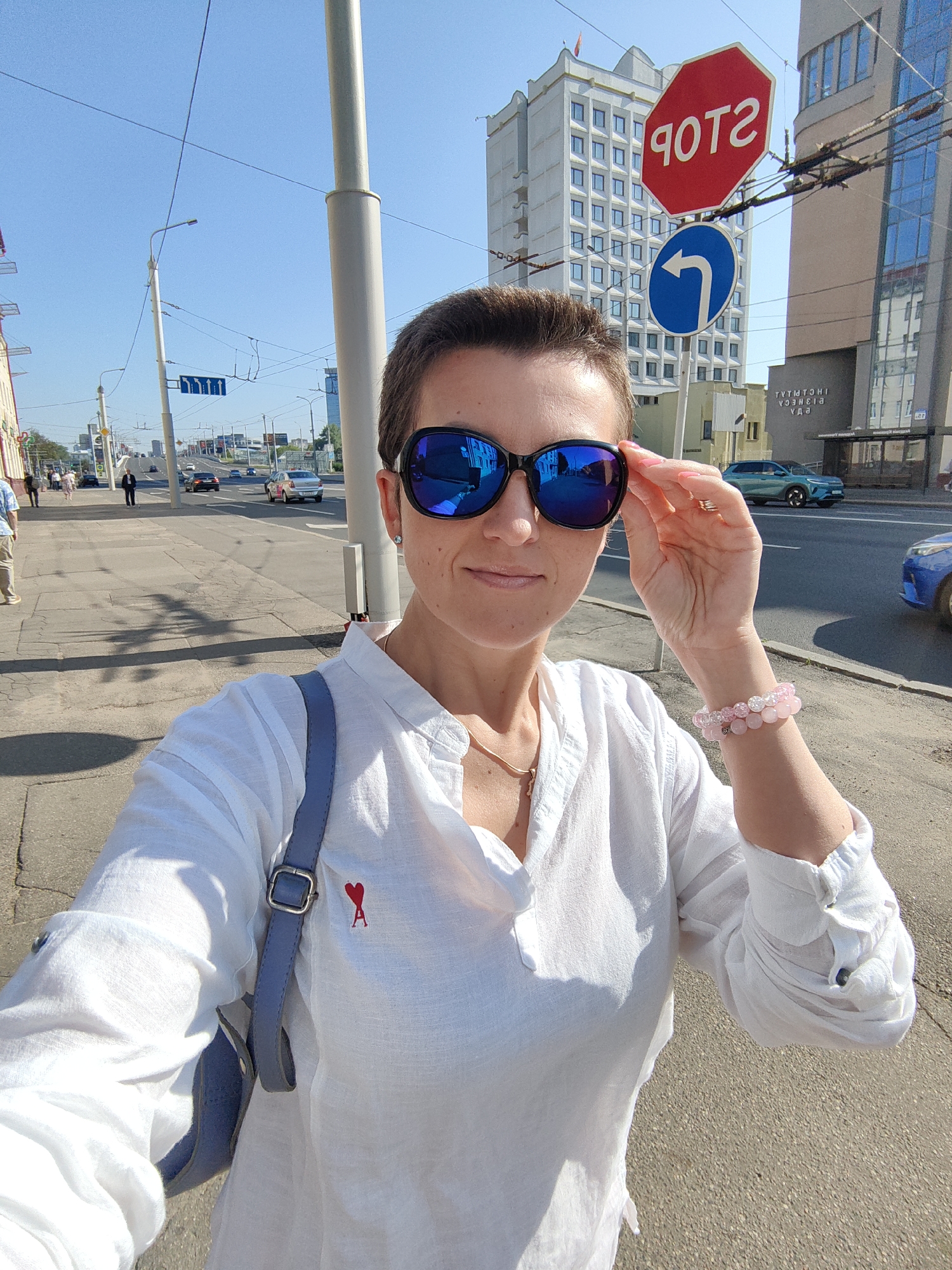 A woman is taking a selfie on a street in Minsk, Belarus. She is wearing sunglasses, a white shirt with a red heart logo, and bracelets. She is standing on a sidewalk with a street and buildings in the background. A "STOP" sign and a turn arrow sign are visible. Cars are driving on the street. The sky is clear and blue.