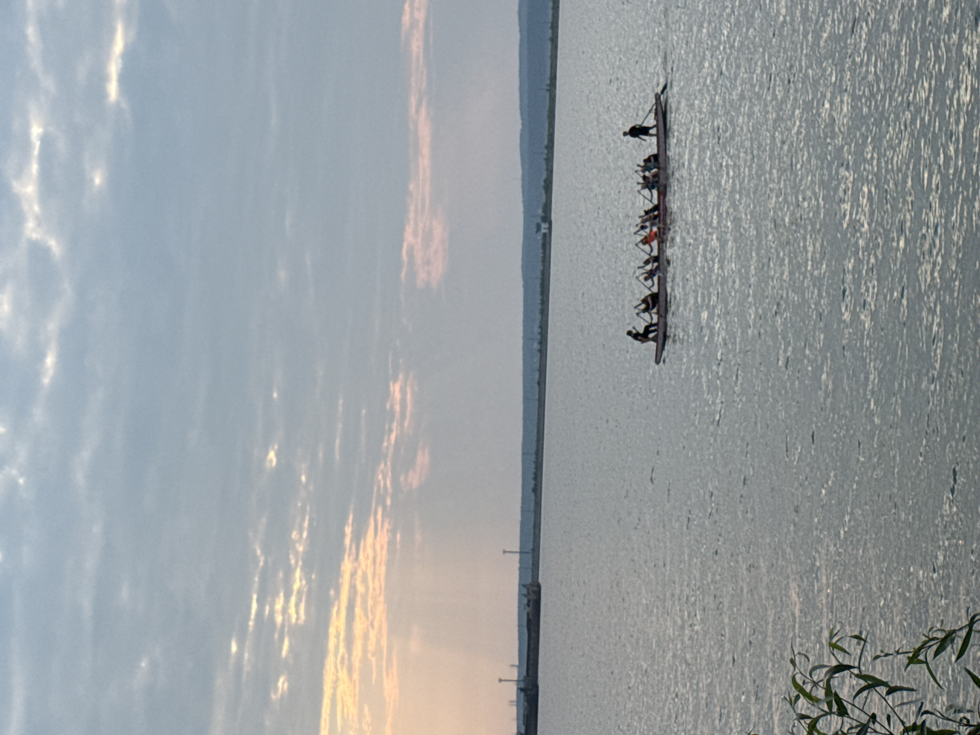 A long, narrow boat with eight people paddling is visible on a body of water under a cloudy sky. The boat appears to be a dragon boat, judging by the number of paddlers and their synchronized movements. The people are dressed in casual clothing and are using paddles to propel the boat forward. In the background, there is a distant shoreline with some structures and trees. The water is calm, with small ripples on its surface. The sky is overcast, with some patches of sunlight breaking through the clouds, creating a soft, diffused light. The overall scene suggests a recreational activity taking place on a body of water, possibly a river or lake, in a natural or semi-urban setting. The image is likely taken during the day, with the soft lighting indicating either early morning or late afternoon.