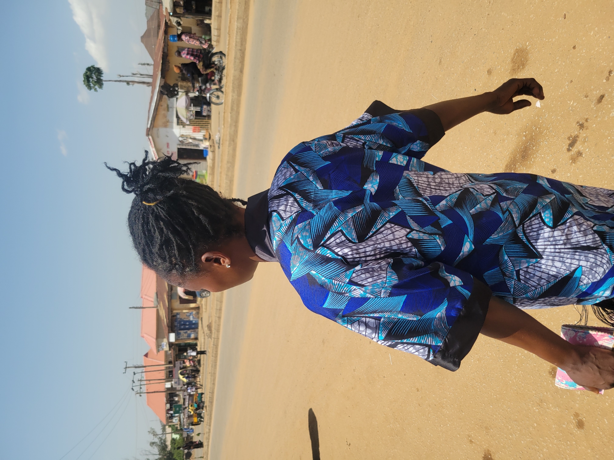 A person with dreadlocks is standing on a paved road, possibly in Rafin Par, Nigeria, as suggested by the presence of buildings and people typical of an urban African setting. The individual is wearing a blue patterned top with black trim. Their right arm is extended, holding a small, white object. In the background, there are buildings, people, and parked motorcycles. The environment suggests a daytime setting, with a clear blue sky overhead.