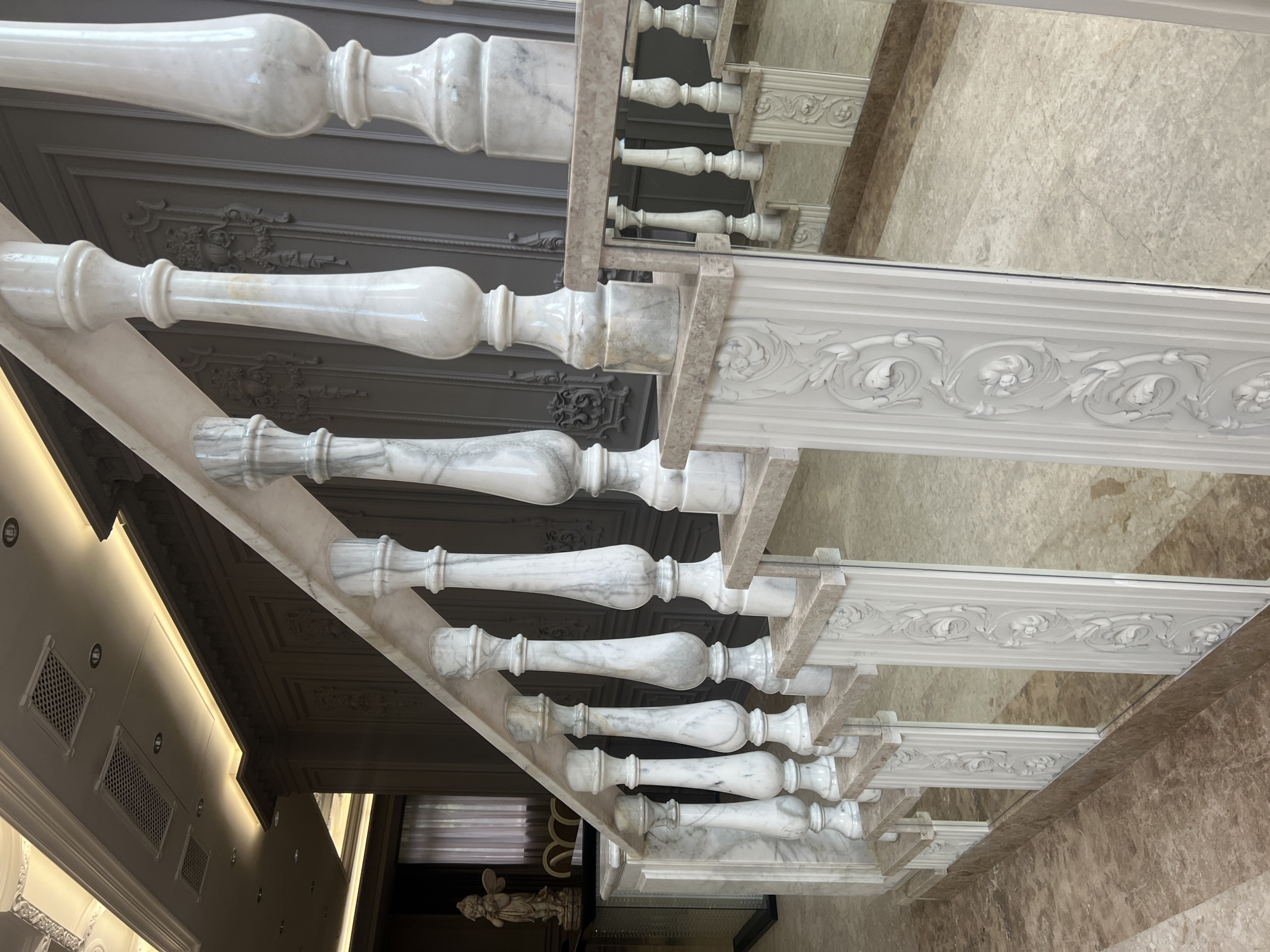 This media file captures an opulent interior architectural detail, likely a grand staircase or a balustrade within a luxurious building in Yerevan, Armenia. The primary subject is an ornate balustrade crafted from white and grey veined marble. It features numerous elegantly turned balusters, each with a distinct, classic profile, supporting a smooth marble handrail. Interspersed along the balustrade are larger, rectangular newel posts, which are intricately carved with floral or scrollwork motifs, emphasizing a classical design aesthetic.
Below the balustrade, a highly polished, speckled marble floor reflects the structure above with remarkable clarity, creating an illusion of extended space and adding to the grandeur of the scene. The reflections mirror the balustrade, the coffered ceiling, and other architectural details.
Above the balustrade, a dark, coffered ceiling with decorative moldings is visible, further contributing to the elaborate interior design. To the side, sections of a wall are shown with integrated modern elements like linear recessed lighting strips and gridded vents, contrasting slightly with the classical style of the balustrade. A small, white, classical statue, possibly depicting an angel or cherub, is partially visible in the lower left corner, adding to the decorative elements.
The setting is static, with no people or activities taking place, allowing full appreciation of the detailed craftsmanship and luxurious materials. The lighting suggests a bright interior, possibly daytime or well-lit by artificial sources. The overall atmosphere conveys a sense of elegance, sophistication, and timeless luxury. No text is visible in the image.