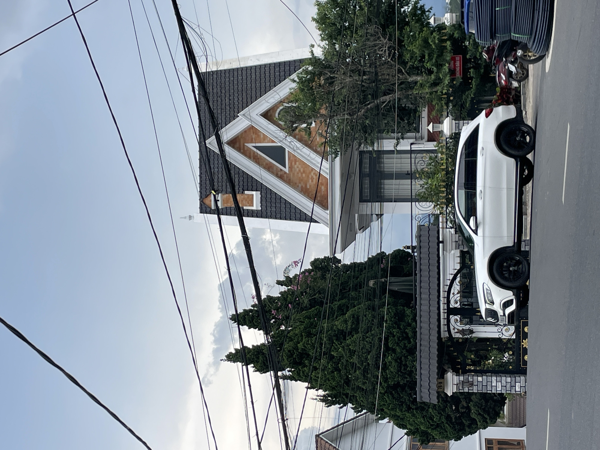 The image depicts a street scene in Phường Xuân Hương - Đà Lạt, Vietnam. A white Mercedes-Benz SUV is parked in front of a gated entrance to a property. Behind the car and gate, there is a multi-story house with a distinctive steeply pitched roof. The property is surrounded by lush green trees. Overhead, a complex network of electrical wires is visible against a cloudy sky. The street in the foreground is asphalt, with a white line marking the edge. To the right of the SUV, a stack of black coiled pipes and two motorcycles are partially visible.