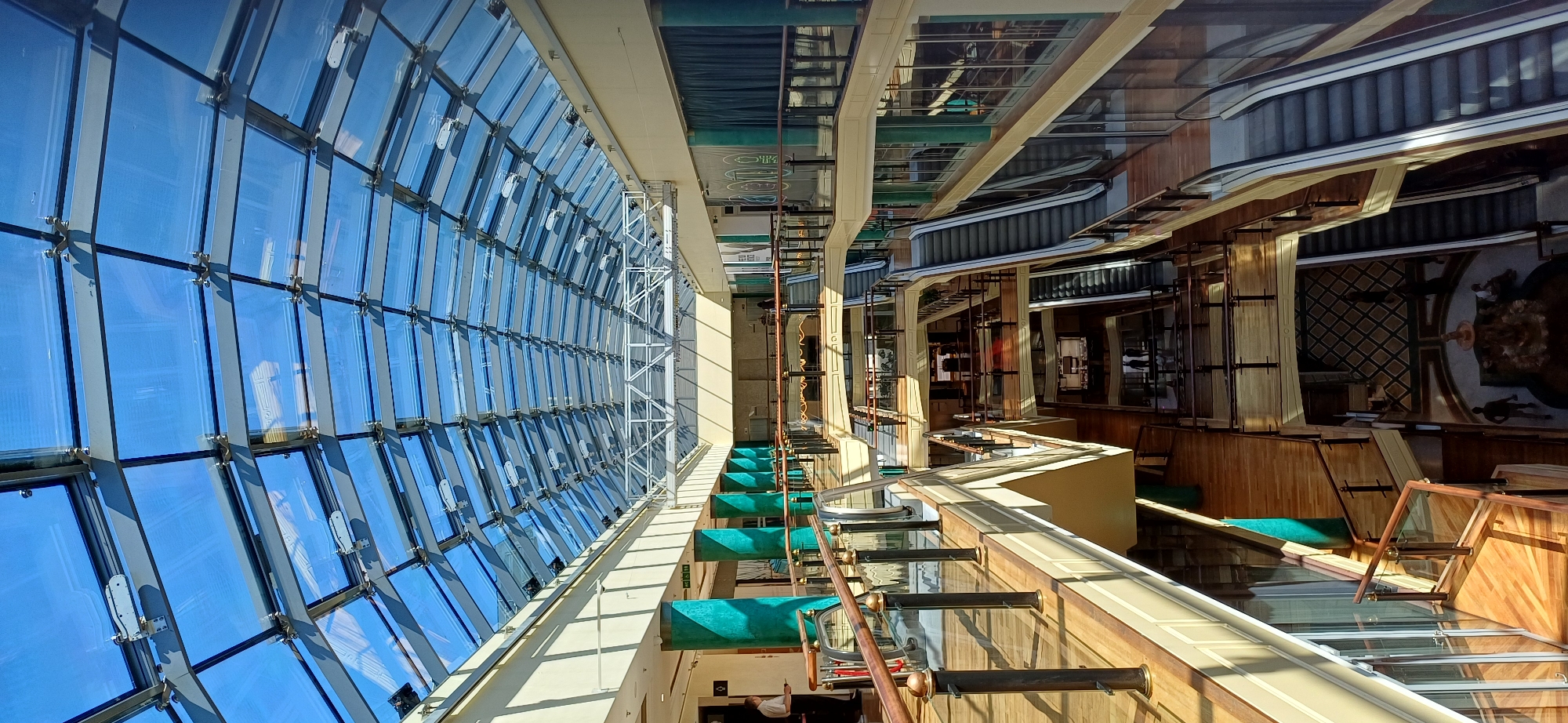 The image shows the interior of a multi-story shopping mall with a glass roof. The mall features several levels with wooden floors and railings. Escalators are visible, connecting the different floors. The ceiling is a complex structure of glass panels supported by metal beams, allowing natural light to filter in. Several turquoise-colored columns are present on the first level. In the foreground, a person in a white shirt is standing near a railing. On the lower levels, a few other people can be seen walking and standing. The overall impression is one of a bright and spacious commercial interior.