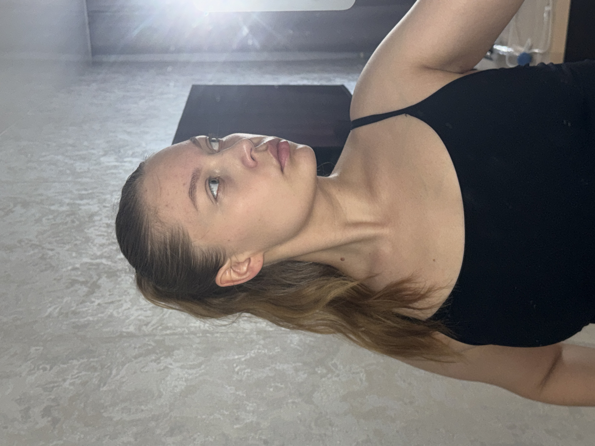 The image displays a young woman lying on a light-colored, textured floor, oriented horizontally across the frame. Her head is positioned on the left side, and her body extends towards the right. She has light skin, light-colored eyes (appearing blue or grey), and brown hair styled with some strands pulled back from her face and longer, wavy sections resting against the floor. A small mole is visible on her neck. She is wearing a black spaghetti strap top.
Her face is seen in profile, looking upwards and slightly to the left of the frame, with a neutral expression. The background features the continuation of the light-colored, mottled floor. In the upper right quadrant of the image (relative to the original orientation), a dark, rectangular object, possibly a low table or piece of furniture, is visible. A strong, bright light source is present at the top edge of the frame, creating a significant glare and lens flare that illuminates the scene. There is no visible text in the image. The depicted scene is an indoor setting. The location context provided is Chaikovsky, Russia.