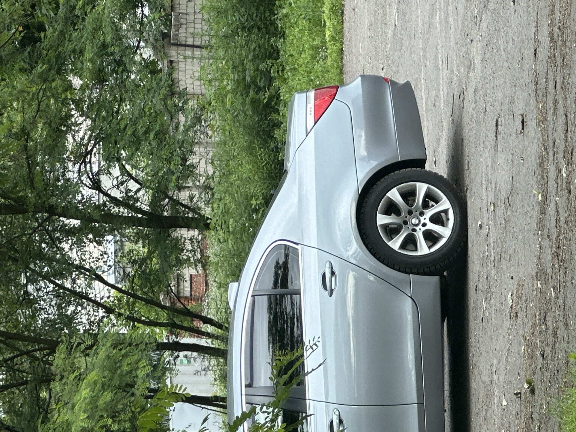 A silver sedan is parked on a gravel surface in front of a brick wall and lush green foliage. The car is viewed from the rear passenger side. The vehicle has tinted windows, a shark fin antenna on the roof, and alloy wheels. The background consists of trees, bushes, and a portion of a brick structure. The lighting suggests it is daytime, possibly overcast. There are no people or other vehicles visible in the frame.