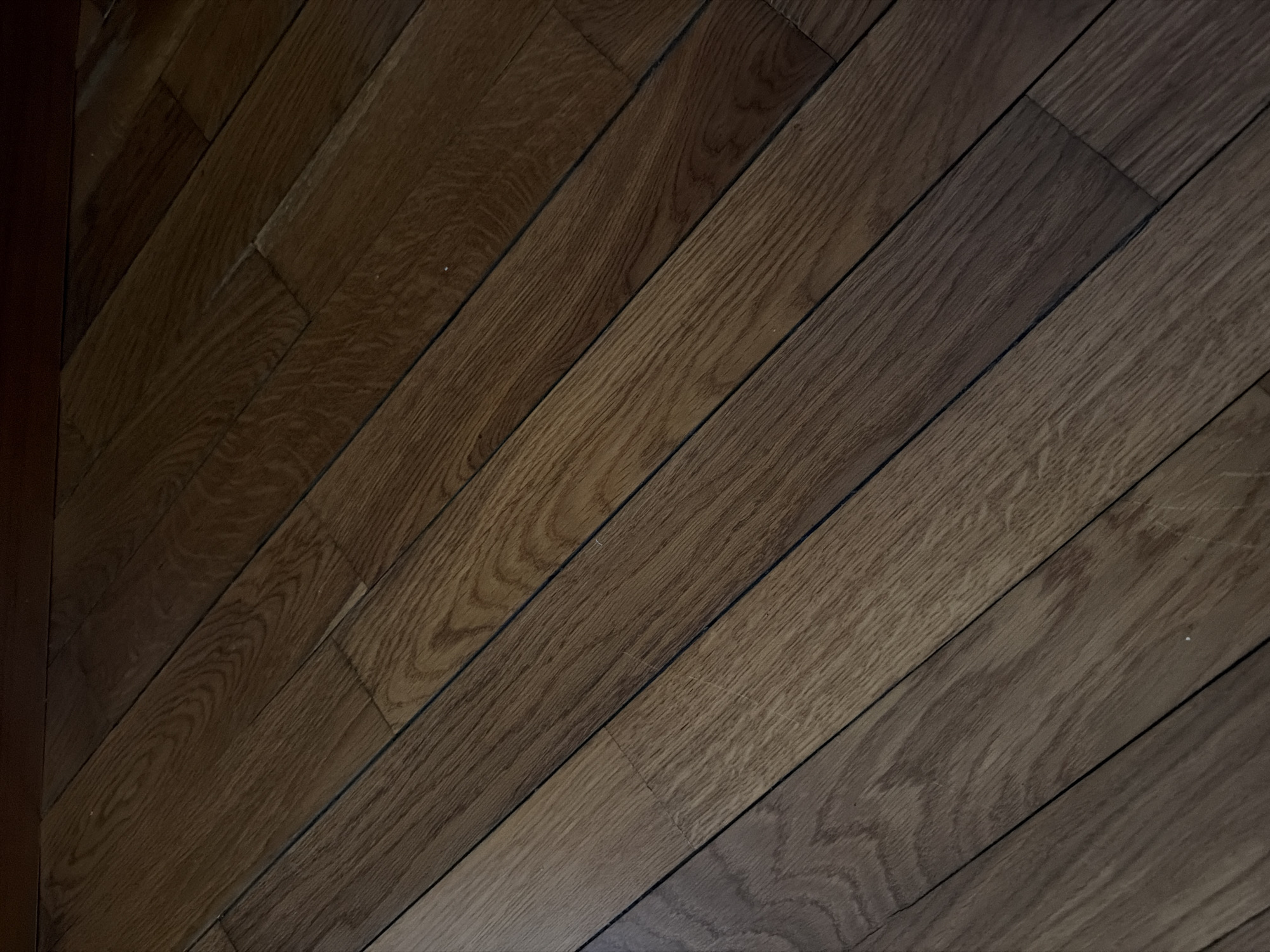 The image depicts a close-up, diagonal view of a wooden floor. The floor appears to be made of planks with a rich, dark brown stain and a visible wood grain pattern. The planks are laid in a pattern that runs from the bottom left to the top right of the frame. The lighting suggests an indoor environment, possibly with overhead light casting subtle shadows. There are no people or discernible objects other than the floor itself. The image does not contain any visual information that would allow for the identification of a specific location within the city of Khmelnytskyi, Ukraine, or any other city.