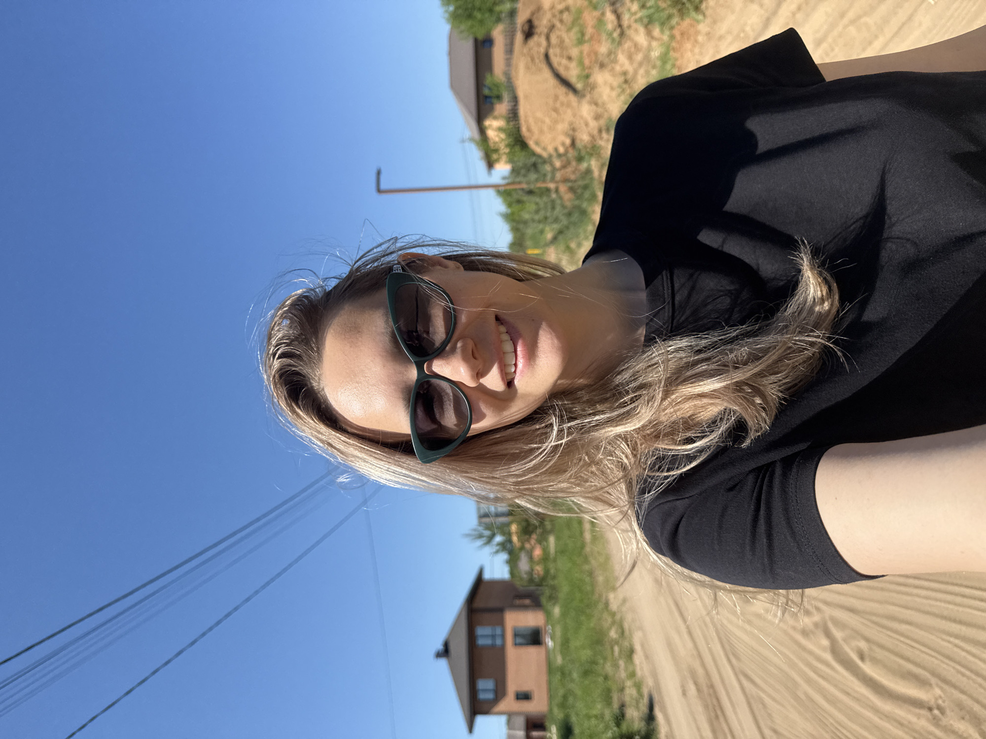 A blonde woman wearing dark sunglasses and a black top smiles into the camera. The photograph appears to be a selfie, taken outdoors on a bright, sunny day. The location seems to be a rural area within the city of Chaikovsky, Russia, as evidenced by a dirt road visible in the foreground and background, a two-story house with a brown roof and facade in the distance, power lines stretching across the clear blue sky, and a sandy hillside partially visible in the upper part of the frame.