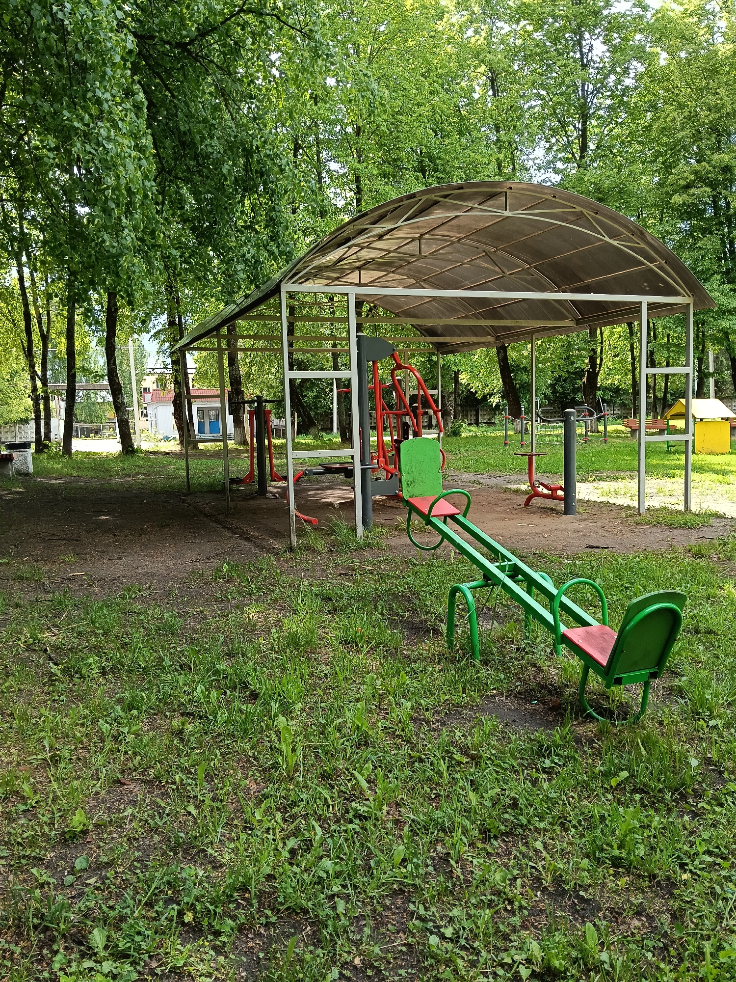 The image shows an outdoor workout area in the city of Красный Маяк, Russia. In the foreground, there is a green and red metal seesaw in a grassy area. Behind the seesaw, there is a covered structure with a metal frame and transparent roof. Under the structure, there are various pieces of outdoor exercise equipment, including a red lat pulldown machine. The ground under the structure is mostly dirt. In the background, there are trees, and a building with a blue door can be seen in the distance. The weather appears to be overcast.