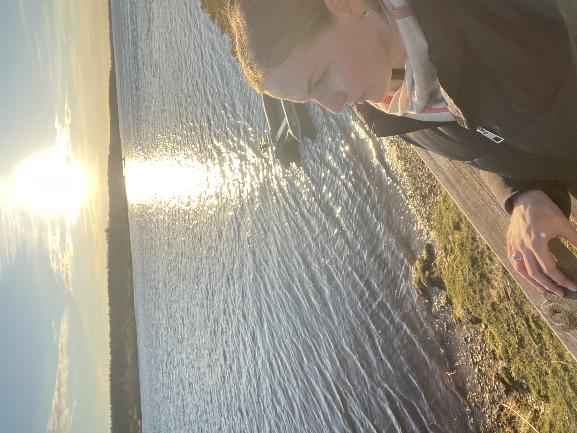 A woman stands facing the water at a location likely in Riga, Latvia. The sun is low on the horizon, reflecting brightly off the water. The woman is wearing a dark jacket and a striped scarf. She is standing next to a wooden railing, with grass and vegetation growing near the water's edge.