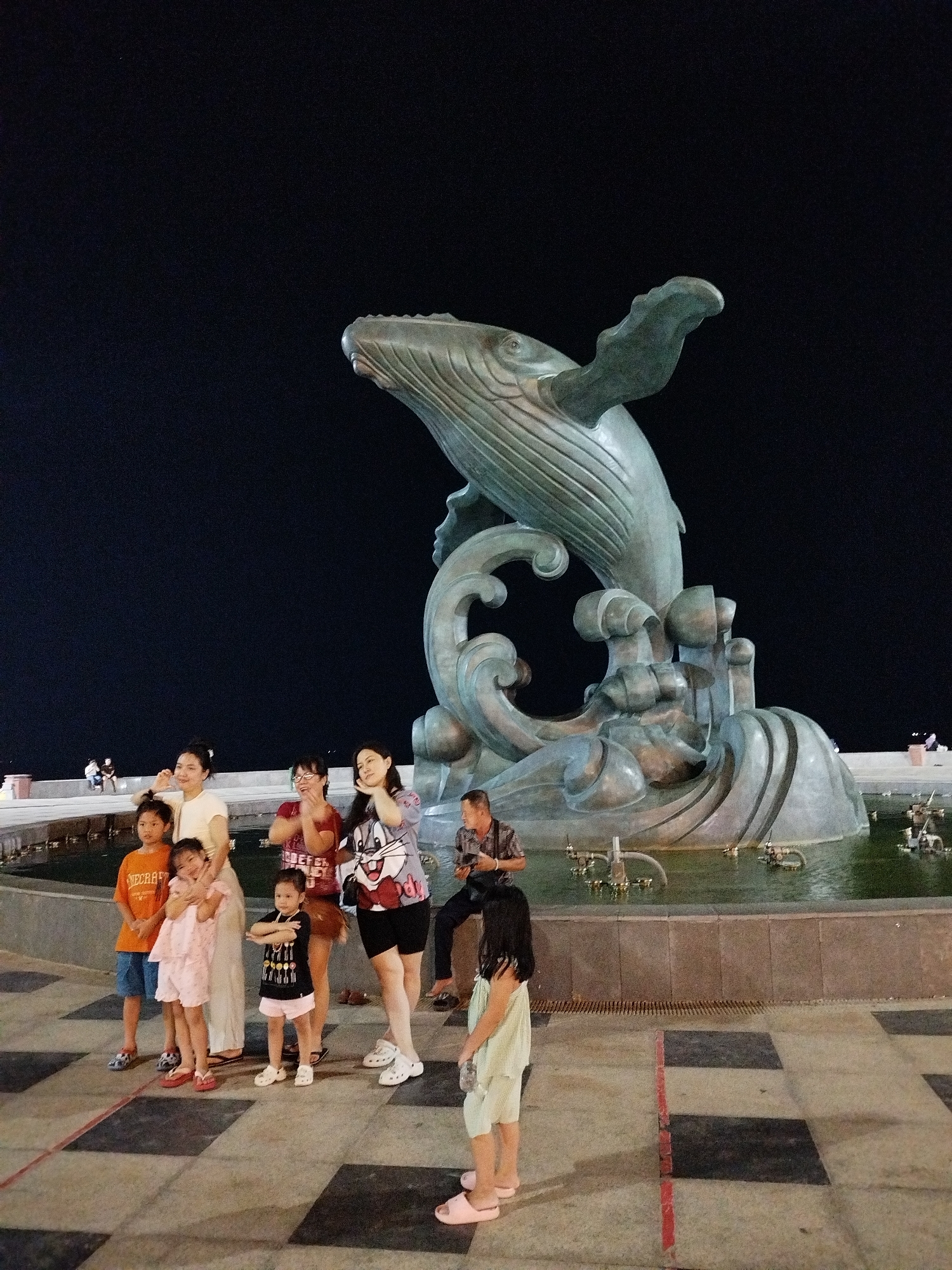 A nighttime scene captures a family standing near a large whale statue in Ho Chi Minh City, Vietnam. The statue, seemingly made of bronze, features a whale leaping from waves, set in a pool with small fountain fixtures. Several people of different ages are grouped near the statue, and one man is sitting behind them, possibly taking photos. The ground around the statue is tiled in a black and tan checkerboard pattern. The background is a dark night sky.