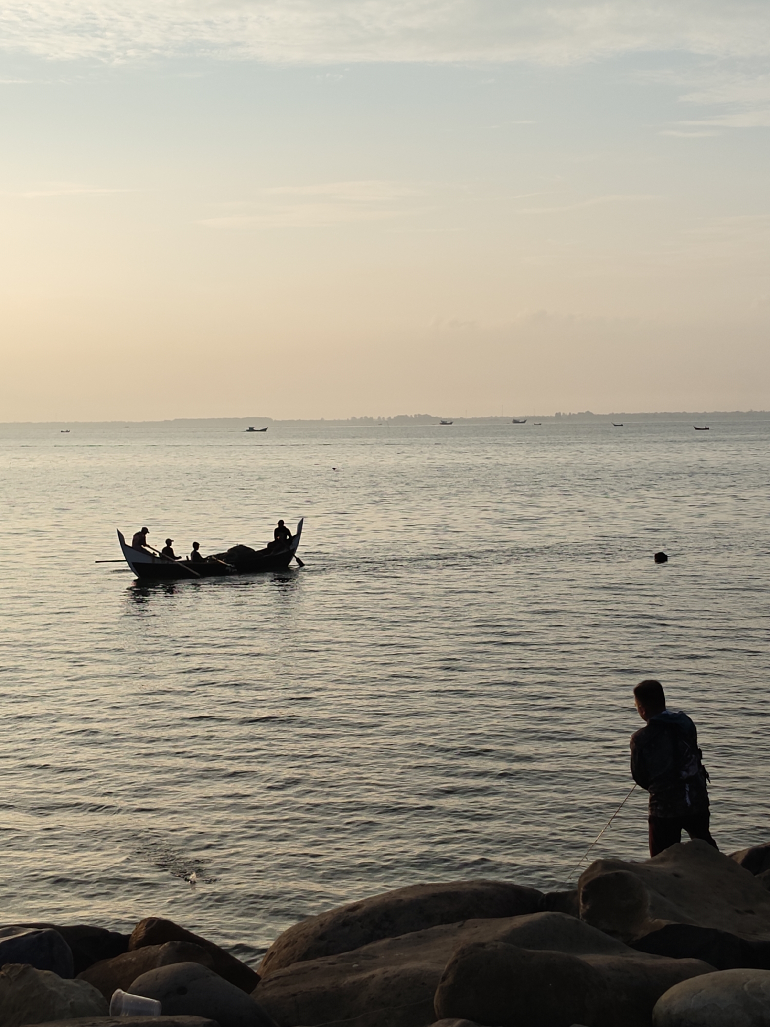 The image depicts a seascape near Lhokseumawe, Indonesia, at what appears to be dusk or dawn, indicated by the soft, warm lighting of the sky. In the foreground, a person stands on a rocky shoreline, possibly fishing with a line cast into the water. In the middle ground, a traditional outrigger canoe carries several individuals. In the background, the horizon line reveals a coastline and a few distant boats. The water is calm with visible ripples.