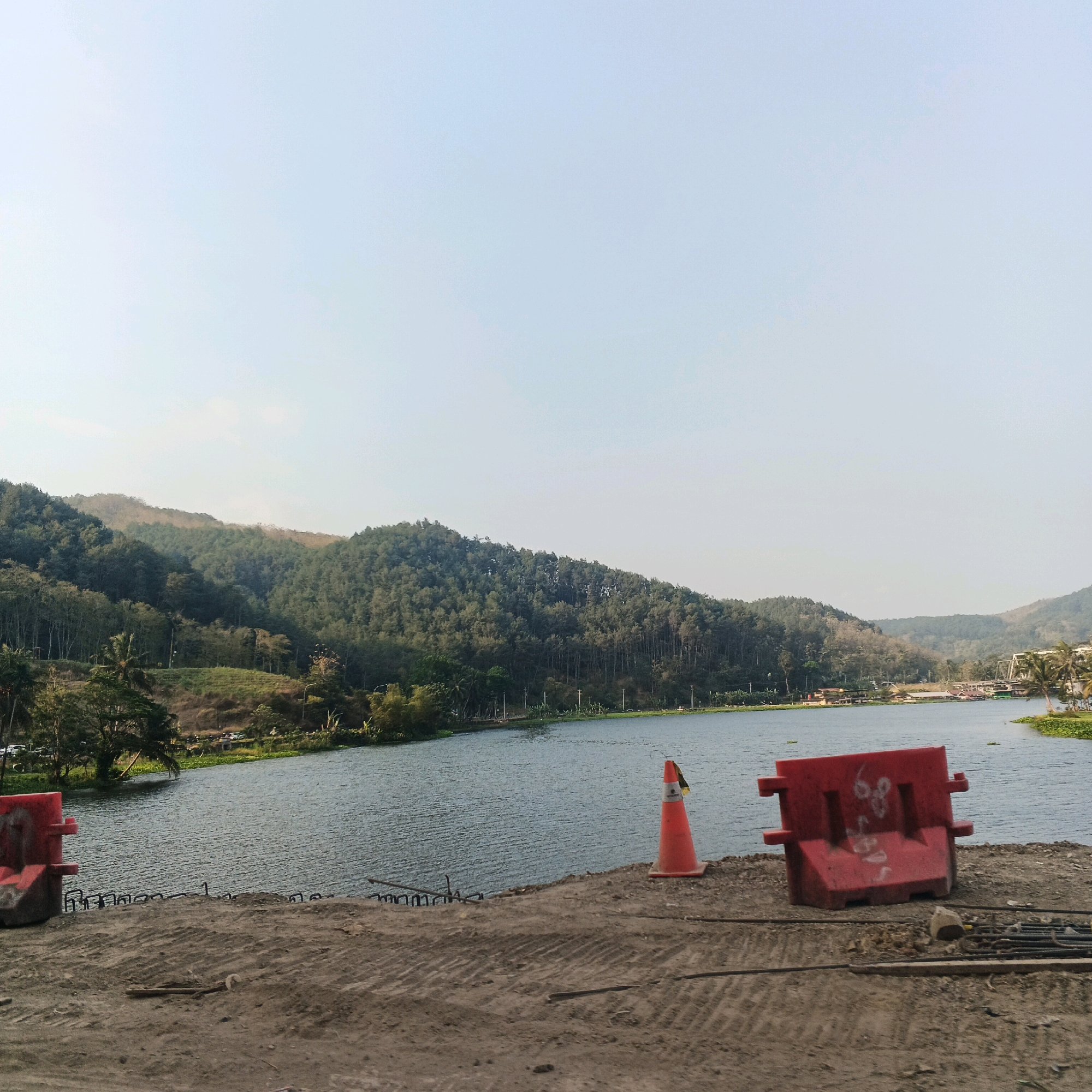 The image depicts a serene lake surrounded by lush green hills and trees, located in Kebasen, Indonesia. In the foreground, construction materials are visible, including rebar and a red barrier. A bright orange traffic cone stands near the edge of the water. The lake is calm, with ripples on its surface. The sky is clear and pale blue. A bridge can be seen in the distance, spanning across the lake, with some buildings and palm trees lining the shore. The overall scene suggests a construction project in progress near a natural landscape.