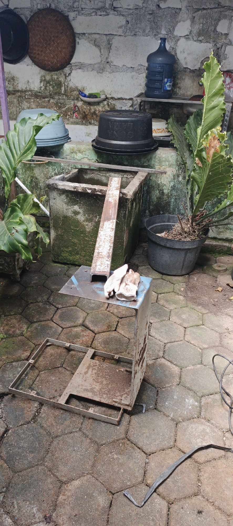 In the city of Moyudan, Indonesia, an outdoor scene depicts a cluttered area adjacent to a stone wall. Dominating the foreground is a rusty metal stand, approximately waist-high, with a smaller metal platform on top where a pair of light-colored work gloves rests. Below this stand, a separate, larger, rectangular metal frame lies on the ground. The ground itself is paved with hexagonal concrete blocks, some of which are covered in moss. Behind the metal stand, a weathered concrete structure, possibly a water tank or planter, is visible. It is also covered in moss and has a metal beam leaning against its side. To the right of this concrete structure, a large potted plant with broad, green leaves stands. Further back, various objects are arranged on shelves against the stone wall, including a large blue water jug, black plastic containers, and a woven basket. The overall environment suggests a utility or storage area, possibly in a residential or workshop setting.
