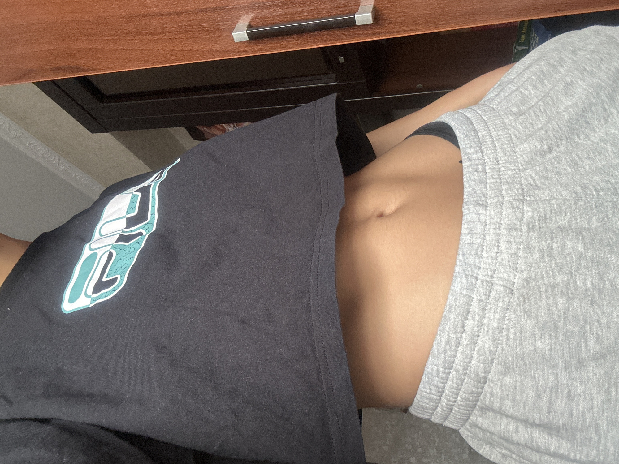 The image captures a close-up, high-angle view of a person's midsection and surrounding indoor environment. The subject's torso is visible, clad in a black t-shirt with a prominent white and teal abstract graphic design on the left side, featuring geometric shapes and a cracked texture. Below the t-shirt, light grey sweat shorts or pants with a distinctive ribbed, elasticated waistband are worn, with a glimpse of a black undergarment visible between the shirt and shorts. The person's navel is clearly visible on their abdomen, which has a light to medium skin tone.
In the background, a wooden surface, possibly a desk or shelf, occupies the top portion of the frame, featuring a reflective silver handle. Beneath this wooden surface is a darker, glossy area, likely another part of the furniture or a shelf. To the upper left, a light-colored, textured wall or surface is discernible. Faint text, including "1990", is barely visible on an object in the shadowy background area, possibly a book on a shelf. The scene is indoors, presenting a static moment without discernible activity or emotion. The overall lighting is soft and even. The location context is Irkutsk, Russia.