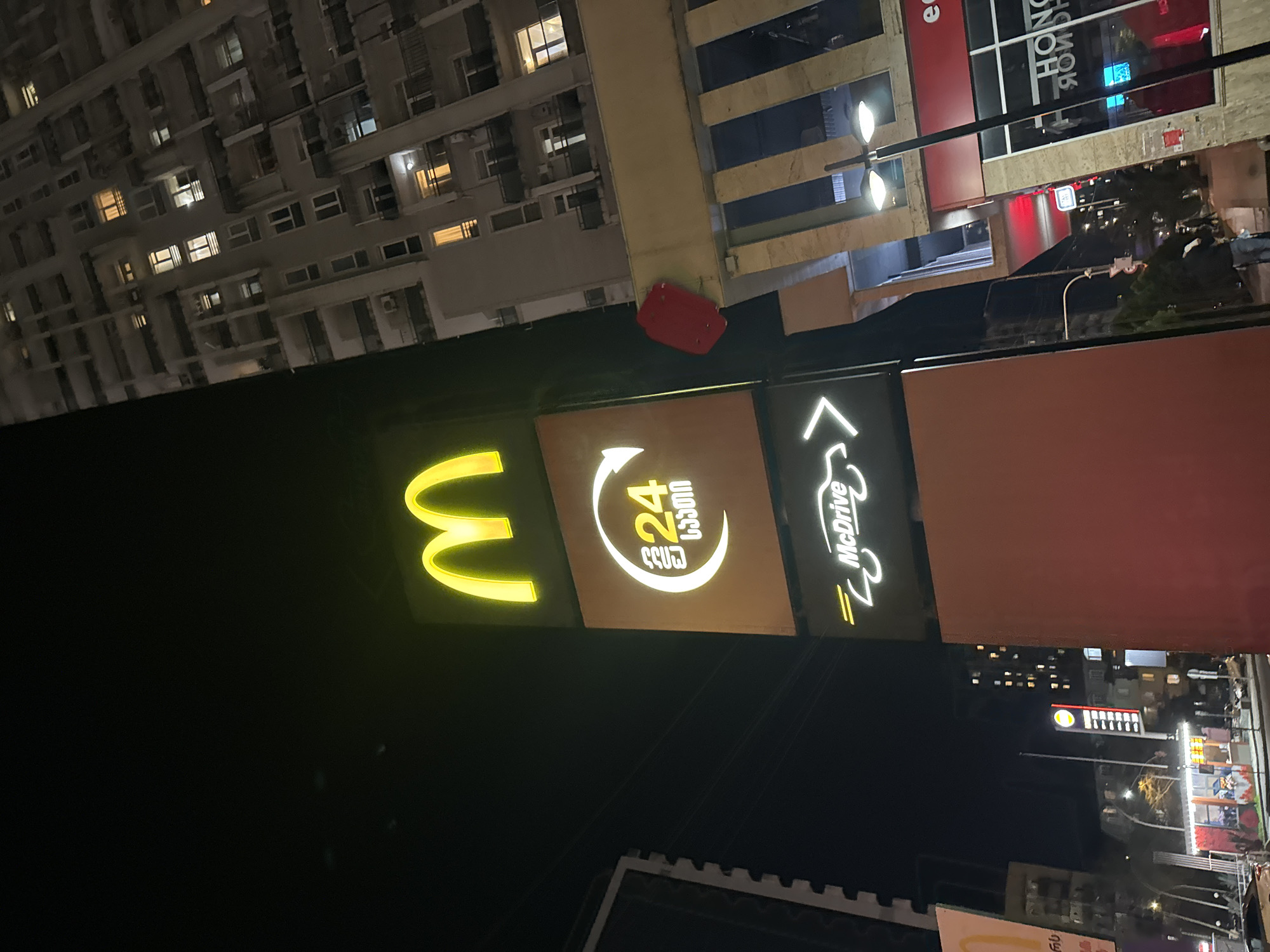 The image is taken at night, showing a McDonald's sign in an urban environment. The sign consists of the iconic golden arches logo, a 24-hour service symbol in what appears to be the Georgian language, and a "McDrive" indication. Tall buildings with lighted windows can be seen in the background, adding to the city environment.