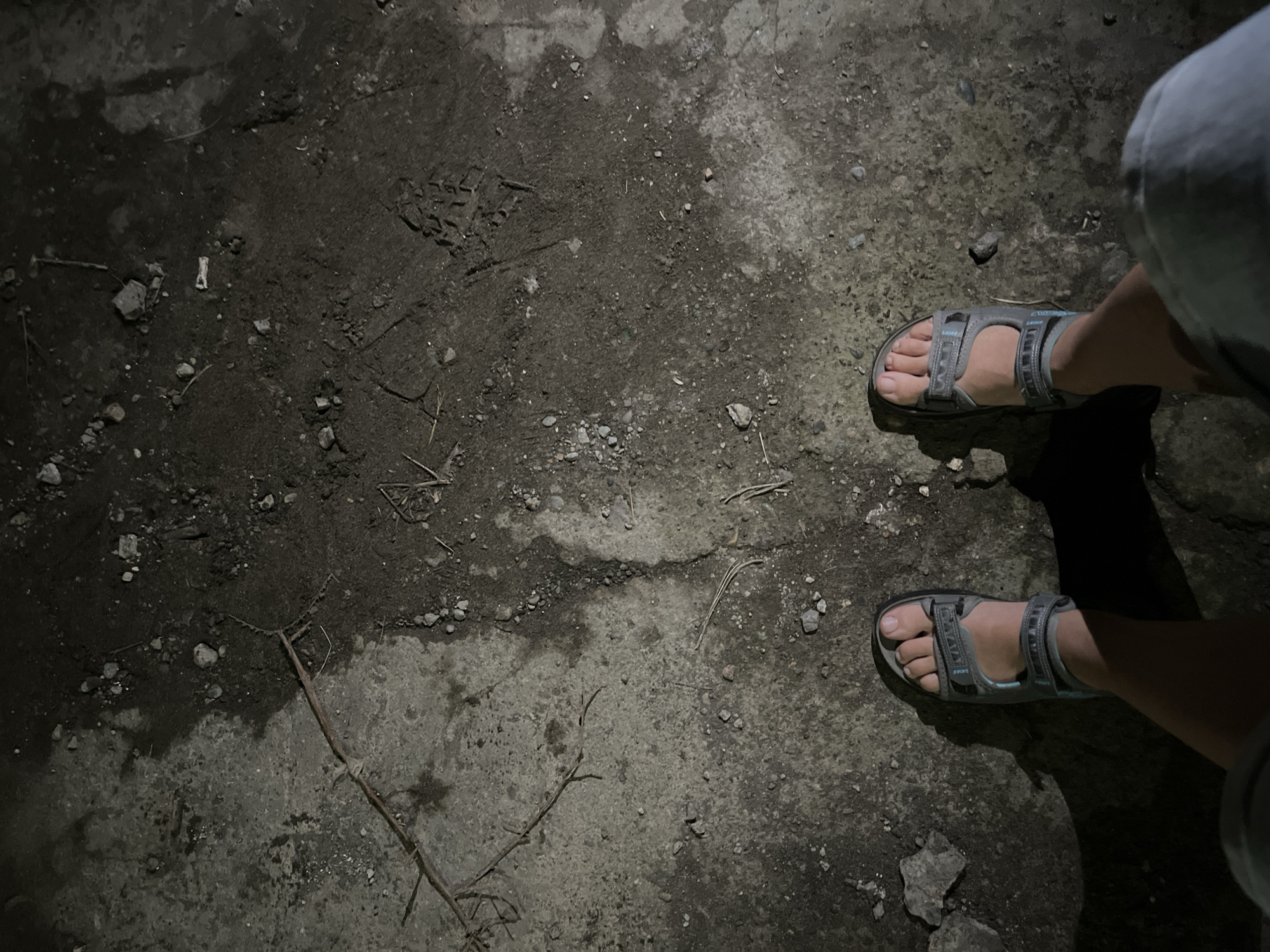 The image shows a first-person perspective of someone standing on a rough, uneven surface, likely a mixture of concrete and dirt. The ground is strewn with small rocks, twigs, and debris. The lighting suggests it is nighttime or a very dimly lit area, casting distinct shadows.
The person is wearing grey and light blue sandals, and appears to be wearing light blue shorts. The sandals are open-toed, revealing bare feet. The overall impression is of an outdoor, possibly unpaved area in a city. Given the context of Poti, Georgia, this could be a street, alleyway, or a less-maintained public space. The scene lacks any explicit identifying markers of Poti, Georgia, beyond the provided location information.