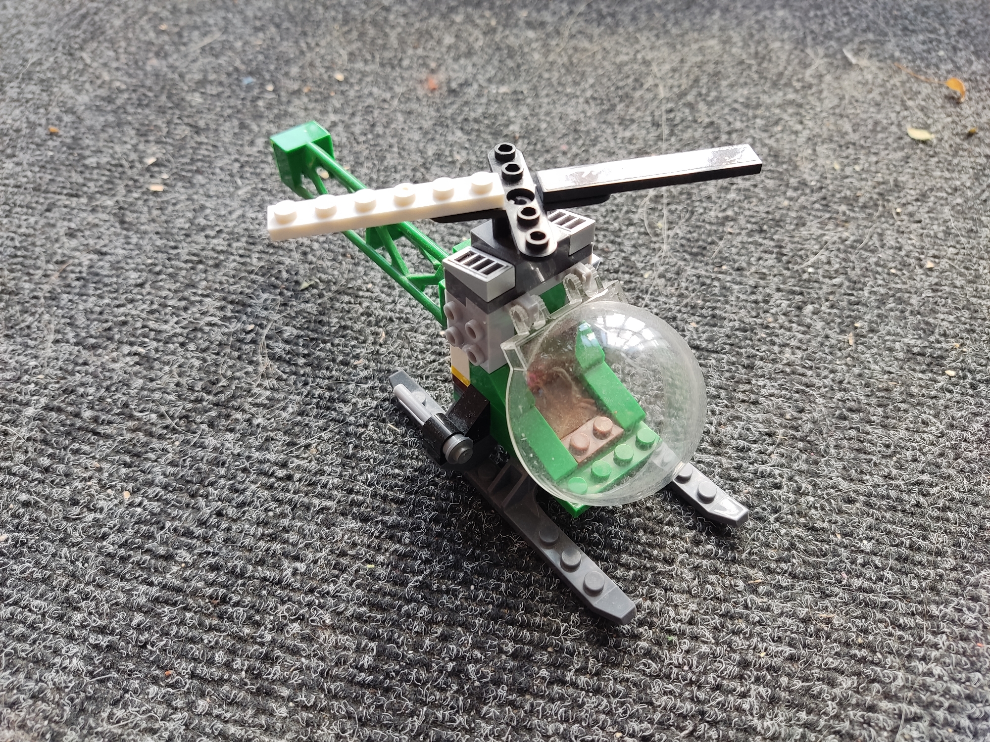 A Lego helicopter is positioned on a dark, textured surface, likely carpet or a similar textile. The helicopter is primarily green and gray, with a clear bubble cockpit. White Lego bricks form part of the rotor assembly. The overall lighting suggests it is daytime, with some shadows cast by the object and its surroundings. The background is out of focus, indicating the camera's focus was on the helicopter. There are no people or discernible actions occurring in the image. The location is unspecified within Бараўлянскі сельскі Савет, Belarus.
