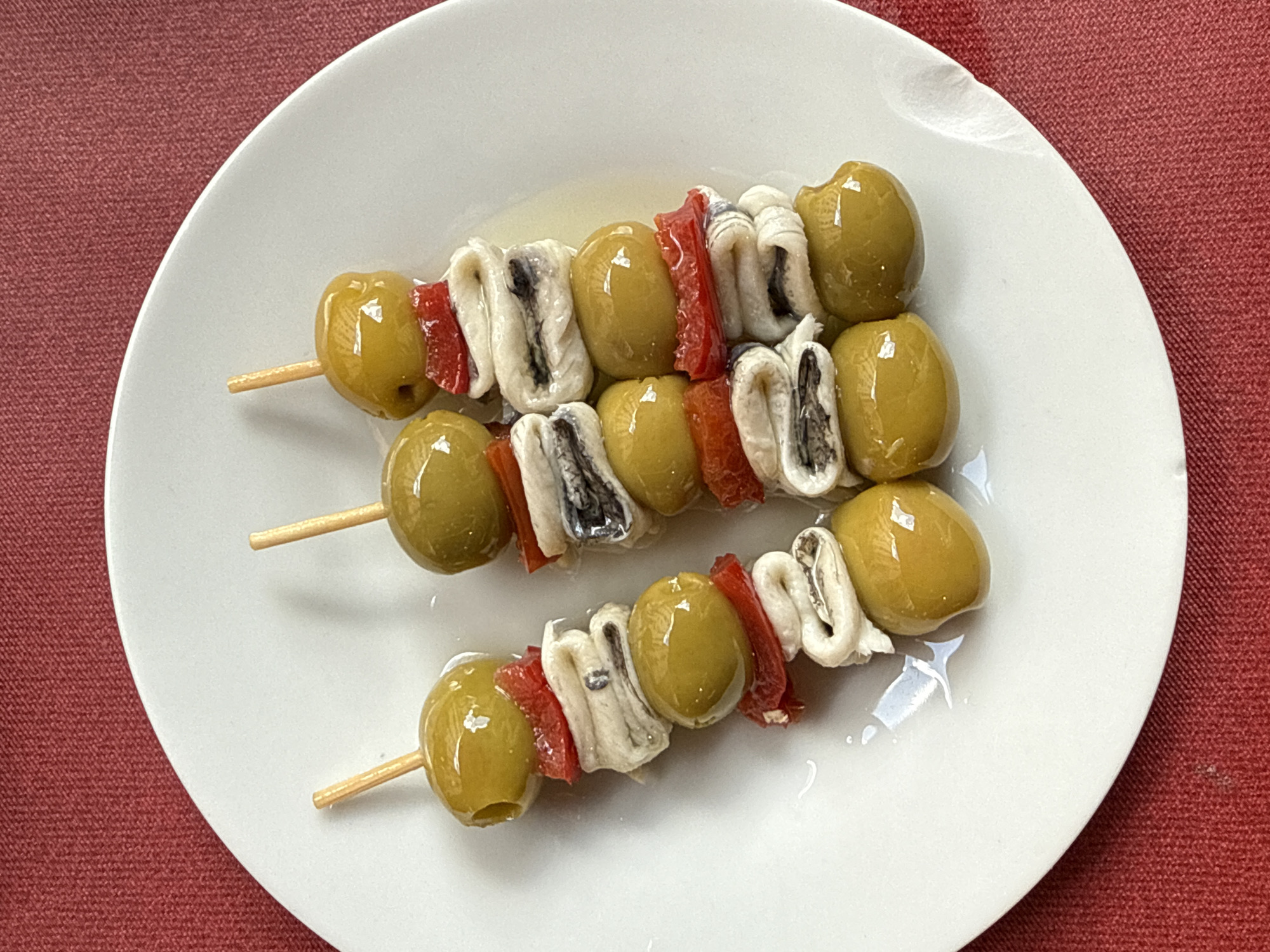The image shows three skewers of "Gilda" tapas on a white plate, set against a red tablecloth. "Gilda" tapas, a typical pintxo from the Basque Country and commonly found in Madrid, consists of olives, anchovies, and pickled peppers threaded on a skewer. The location is most likely in Madrid, Spain, due to the widespread availability of this tapa in the city. The plate appears to have a glossy finish. The skewers are arranged linearly, with the olives and peppers glistening, indicative of being preserved in brine or oil.