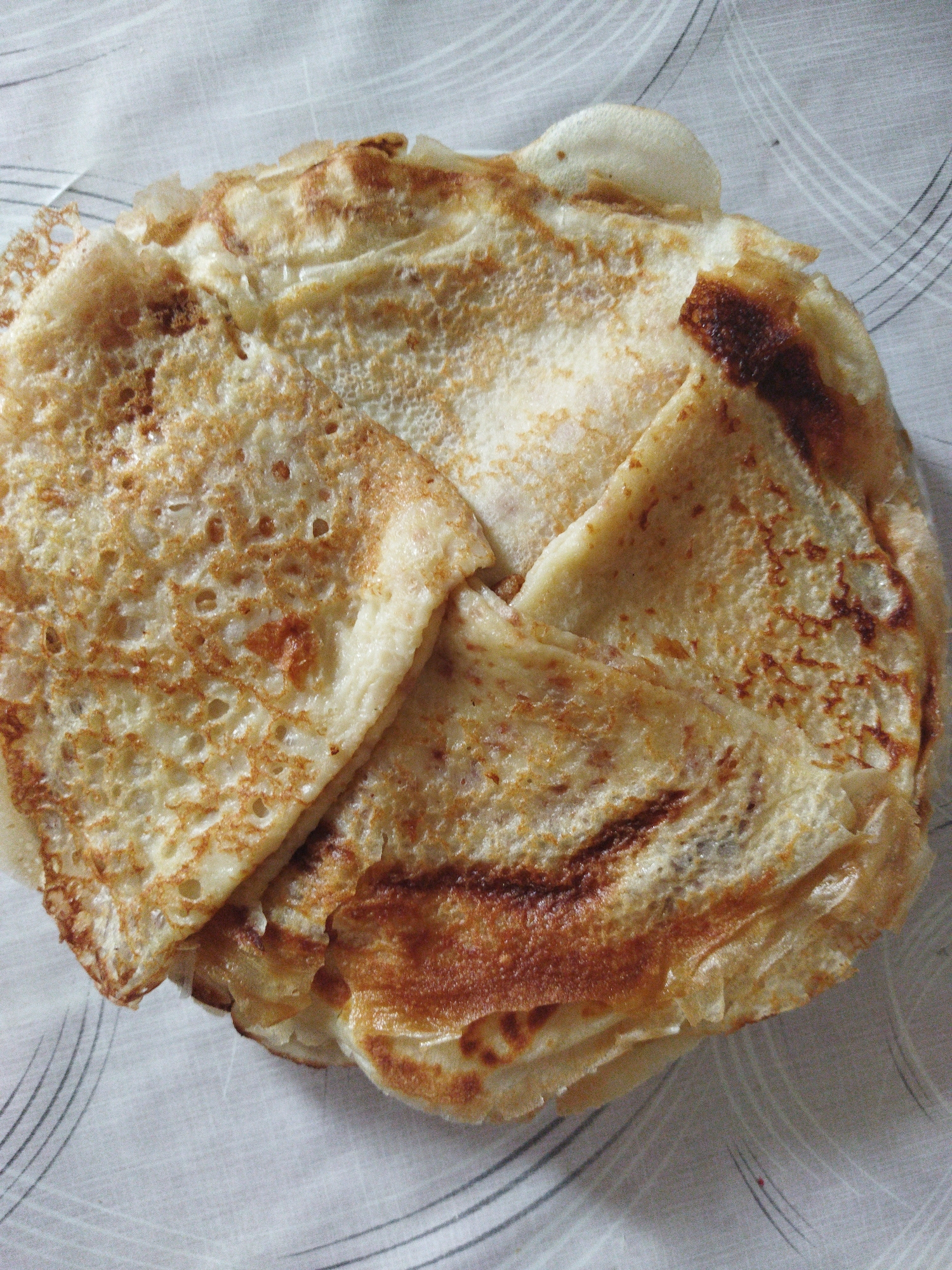 A close-up, overhead shot shows a stack of golden-brown crepes on a white, patterned tablecloth. The crepes are folded and layered, with some appearing slightly crispier than others, showing small bubbles and a slightly irregular texture. The lighting highlights the texture and browning of the crepes. The background is a light-colored tablecloth with faint, swirling grey lines.