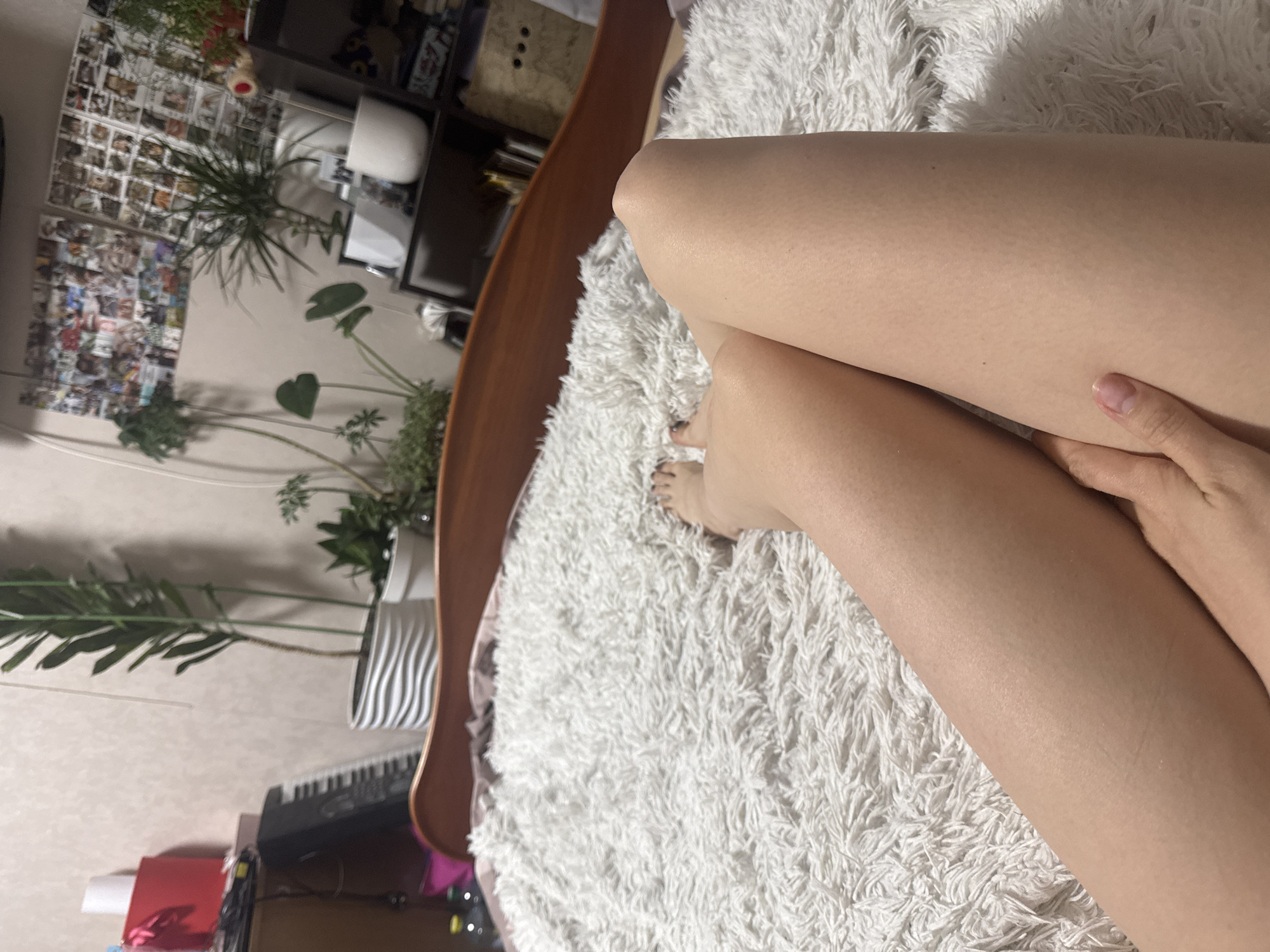 The image captures a person's bare legs, primarily the thighs and lower legs, resting on a fluffy white blanket that covers a bed. One hand is visible, gently touching a thigh. The person's bare feet are partially visible towards the bottom of the frame, with painted toenails. The setting appears to be a bedroom or living area.
In the background, visible behind the bed and rotated due to the camera angle, is a decorated room. A large section of the wall is covered with a collage of numerous small photographs or prints. Several potted plants of varying sizes are arranged around the room, including a tall, leafy one and smaller ones on shelves. Dark wooden furniture, possibly shelves or a bed frame, is visible on the left and bottom. Other objects include what appears to be a musical keyboard on a surface in the lower left background, and a white ceramic item on a shelf above.
The activity depicted is one of relaxation or resting on the bed. The lighting suggests it is daytime, with bright light illuminating the scene, particularly the textured blanket. No discernible text is visible within the image.