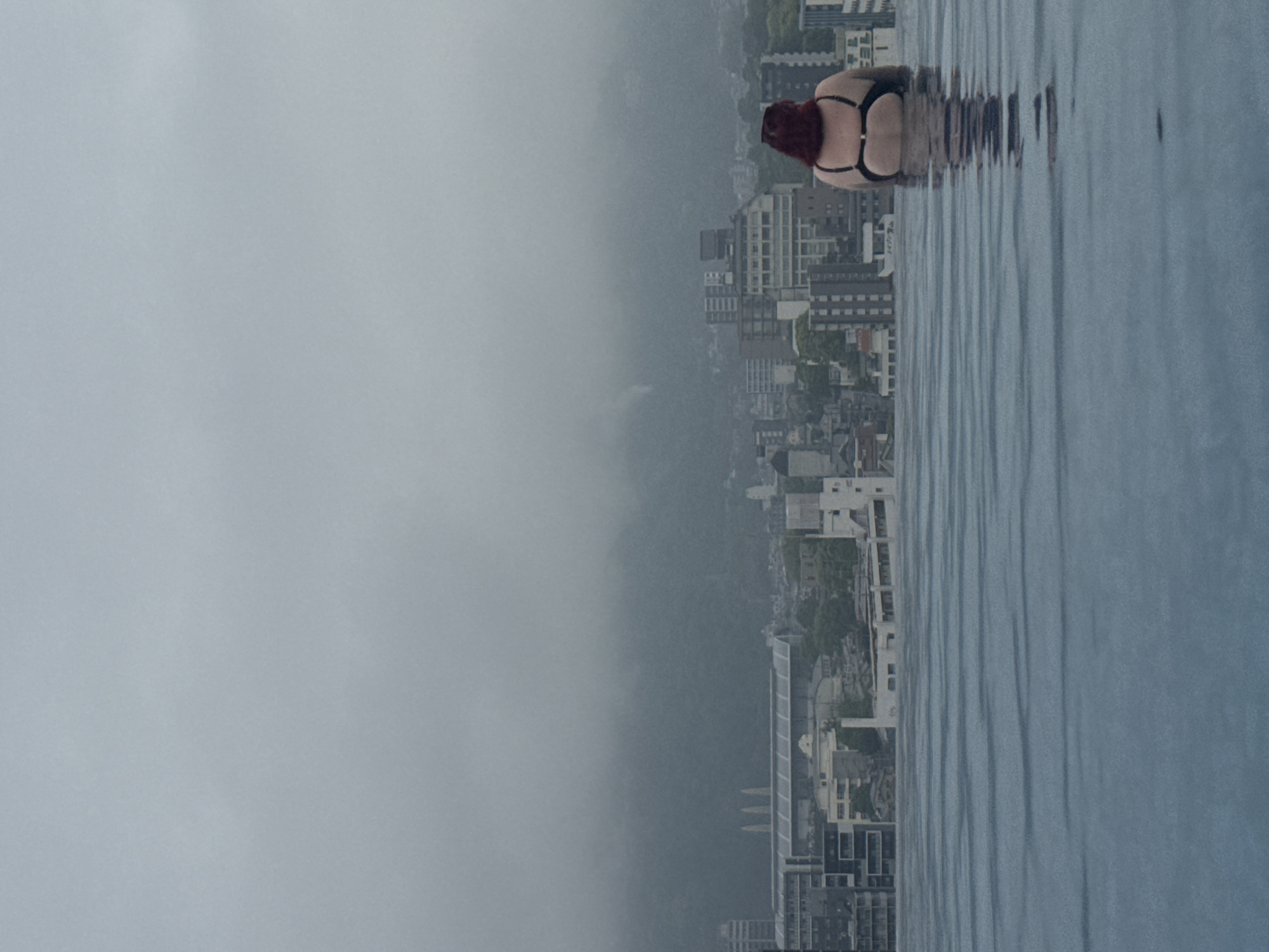 The photograph presents an exterior view likely taken from the ANA InterContinental Beppu Resort & Spa in Beppu, Japan. The perspective is from an elevated position, possibly a rooftop pool or high floor. A woman is visible at the edge of the water, wearing a black bikini. She has auburn or red hair, and her body faces away from the camera, partially submerged in the water.
The background reveals a dense urban landscape with a mix of residential and commercial buildings, including a variety of mid-rise and high-rise structures. The visibility is somewhat obscured by a heavy fog or mist, which reduces the clarity of the distant cityscape. This fog covers a substantial portion of the image, adding to the atmospheric perspective.
The water is calm with slight ripples on the surface. The color is a dull gray/blue reflecting the overcast conditions. The overall ambiance of the scene is subdued and tranquil due to the heavy mist and lack of strong sunlight.