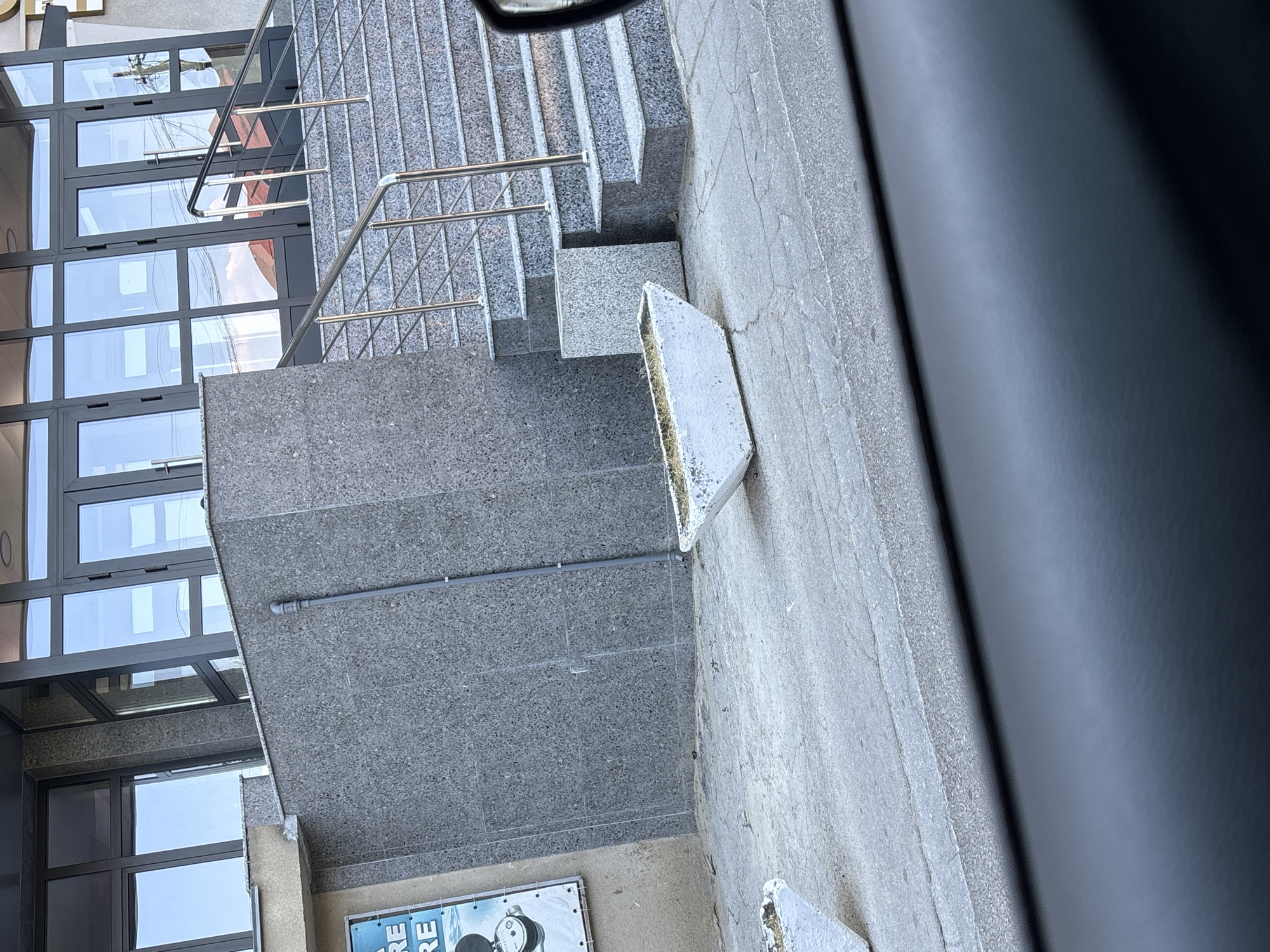 The image depicts an outdoor scene with architectural elements. A concrete staircase with metal railings leads upwards, constructed from speckled grey material. Adjacent to the stairs is a building with large glass windows, reflecting the sky and some reddish-brown structures. A white, rectangular planter with dried grass is positioned near the bottom of the stairs. A metal conduit runs along the grey concrete wall. In the lower portion of the image, a small section of a poster with the text "RE" is visible, along with a graphic resembling a film projector and the word "RE" repeated. The scene appears to be in an urban setting, likely during daylight, with overcast or bright sky conditions suggested by the reflections in the windows. The overall mood is neutral and observational, focusing on the architecture and urban landscape.