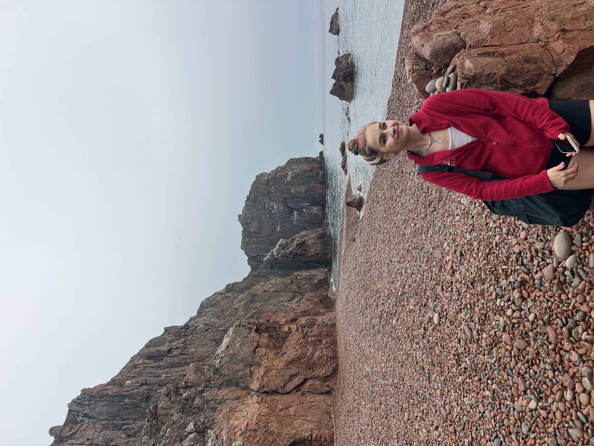 A young woman stands on a pebble beach in Russkiy Island, Russia, against a backdrop of rugged red rock cliffs and the sea under an overcast sky. She wears a red zip-up jacket over a white top, black shorts, and has a backpack over one shoulder. Her hair is pulled back in a bun, and she is holding a smartphone. The beach is made up of small, reddish-brown pebbles, and a large rock formation is visible in the background near the shoreline. The sea is calm with scattered rocks breaking the surface. The sky is a uniform grey.
