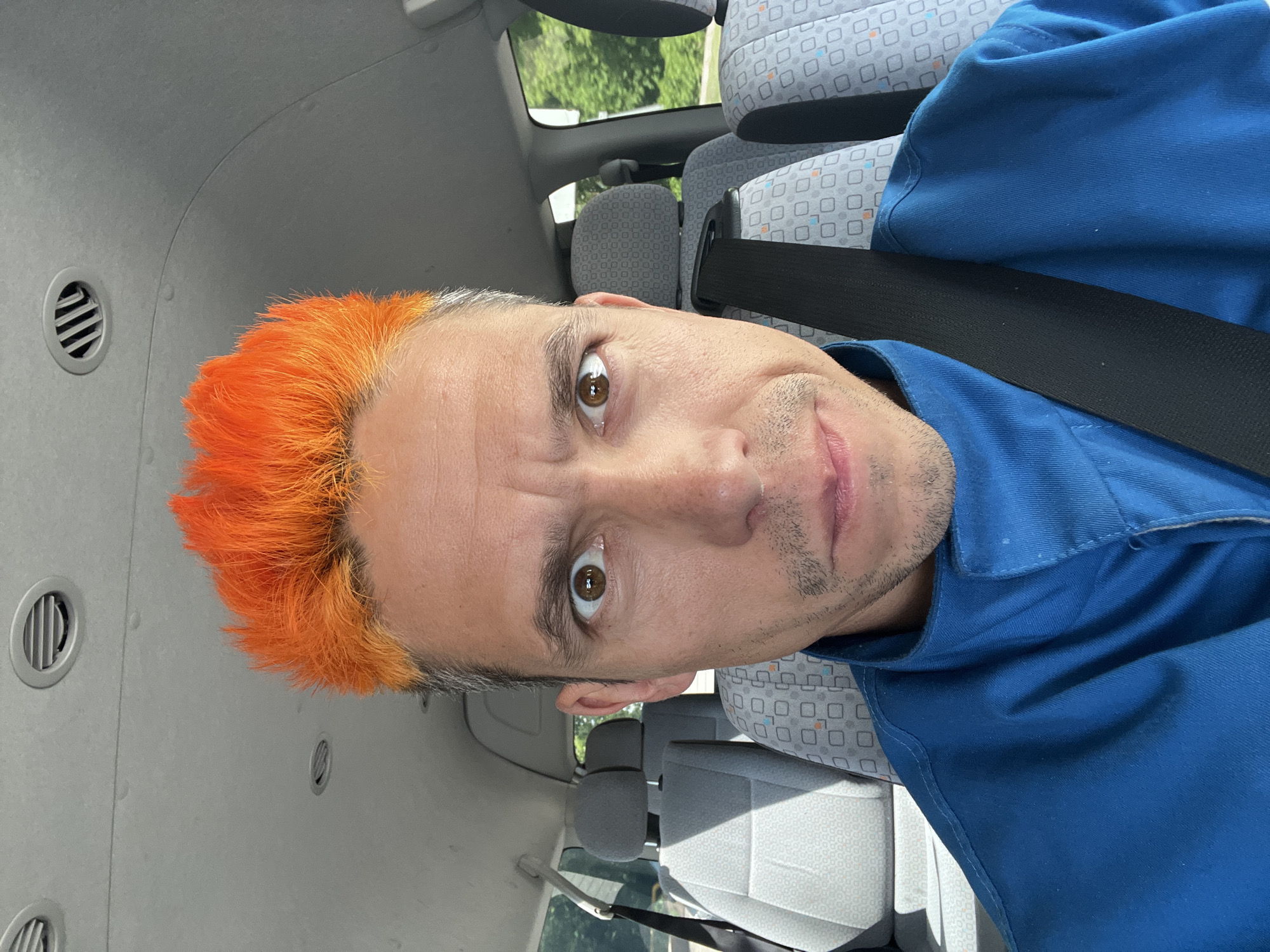 A man with orange hair is wearing a blue shirt and a seatbelt in what appears to be the interior of a vehicle. The seats have a patterned upholstery. The location is unknown.