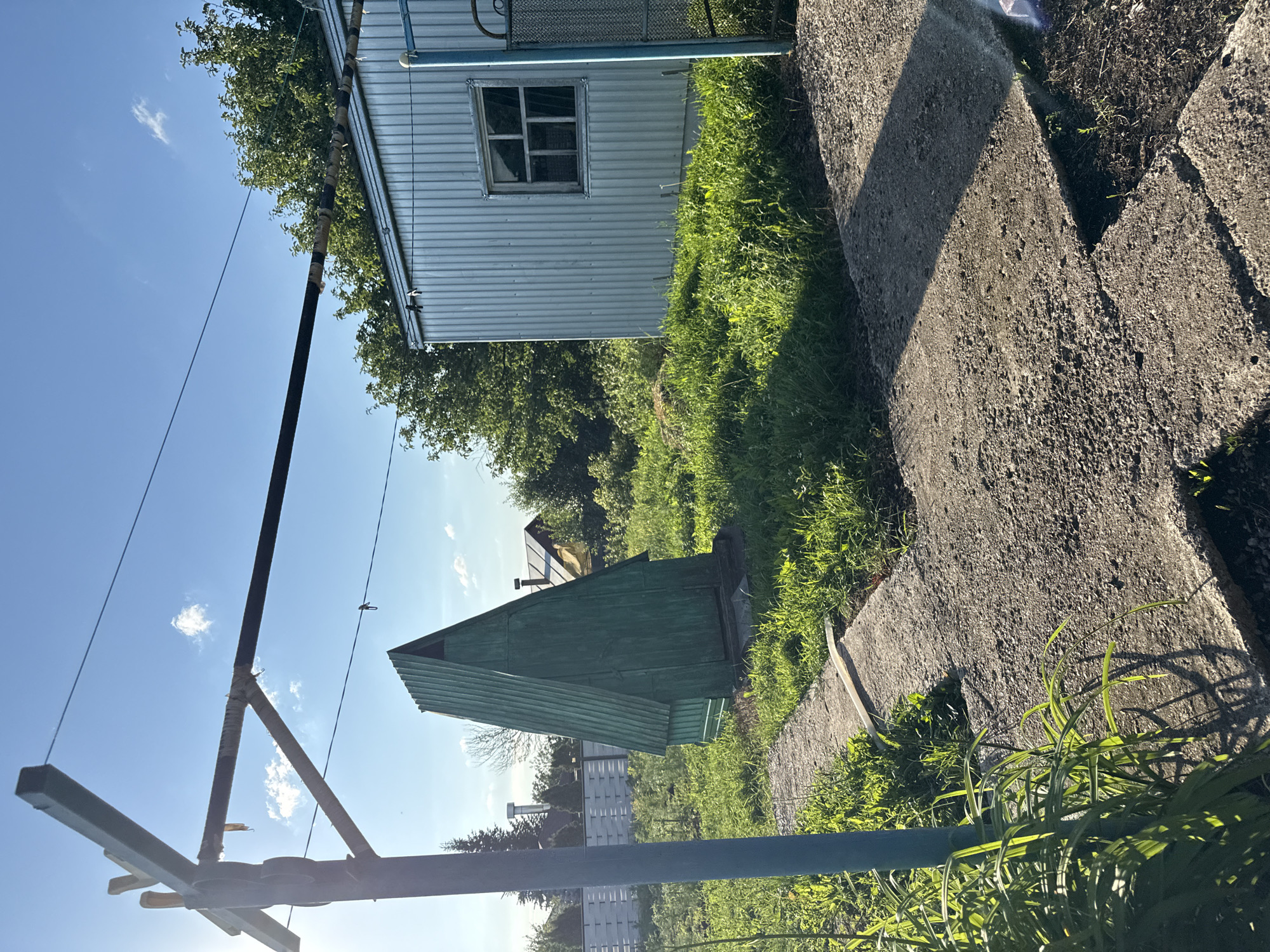 The image captures a rural scene in the village of Verkhneyarkeyevo, Russia, during daylight. In the foreground, a concrete pathway is visible, partially covered by green grass and some leafy plants. To the left, a vertical blue pole supports a wire that extends horizontally across the frame. Behind the pathway, a tall, green, A-frame structure stands prominently in a grassy area. Further back, a corrugated metal building with a small, multi-paned window is situated to the right. Lush green trees and vegetation surround the structures. The sky above is blue with scattered white clouds, and the sun is shining, casting shadows. The overall impression is one of a functional, outdoor space in a somewhat rustic setting.