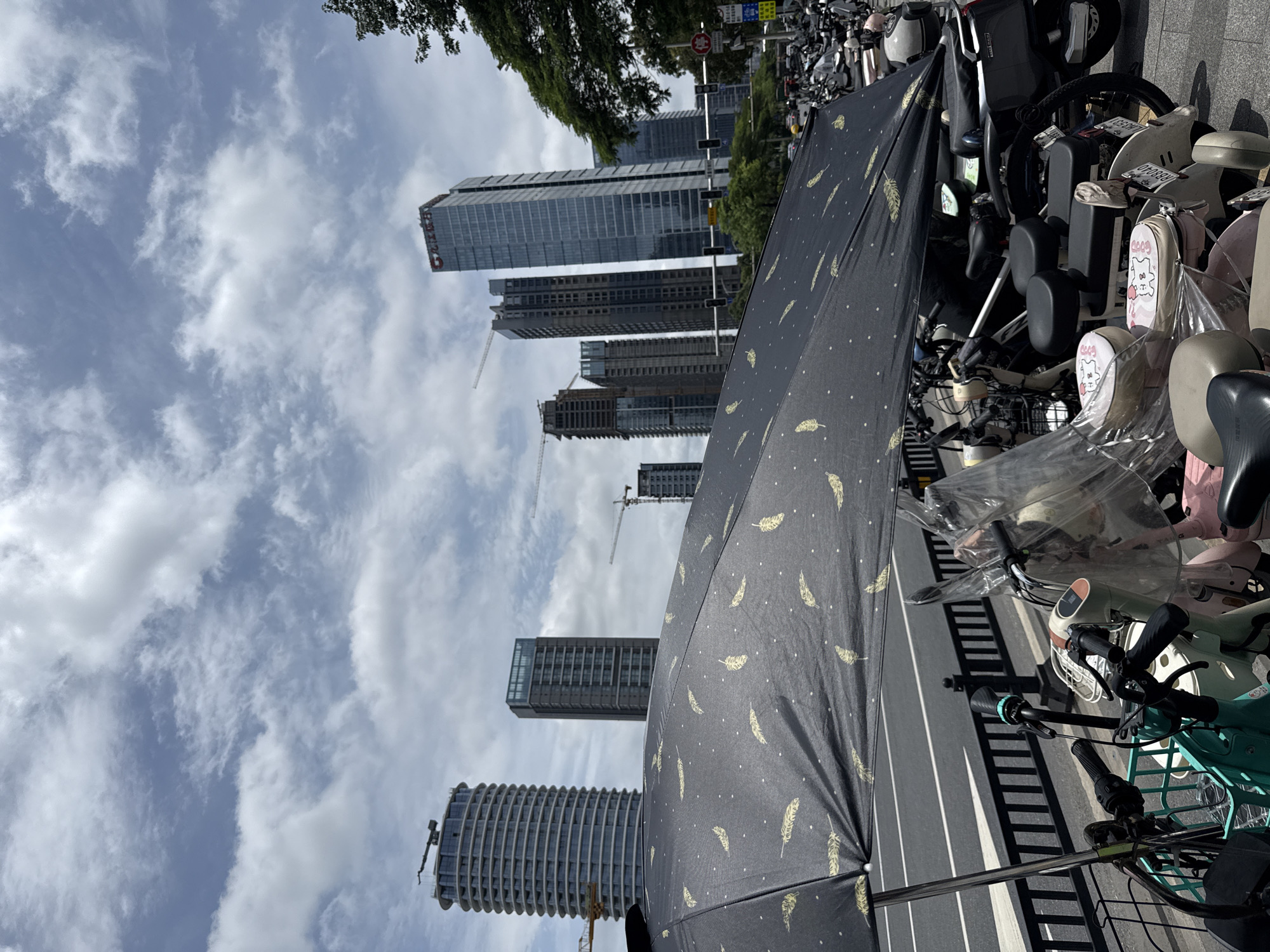 In Tianhe District, China, under a cloudy sky, a black umbrella with a repeating feather pattern is open and angled to cover a row of parked electric scooters and bicycles. The foreground is dominated by the umbrella and the handlebars and seats of the vehicles. Several scooters and bicycles are visible, some covered with clear plastic sheeting. In the background, a modern cityscape rises, featuring several tall, glass-fronted skyscrapers and two active construction cranes. A traffic light is visible in the middle ground, suggesting the scene is near a road.