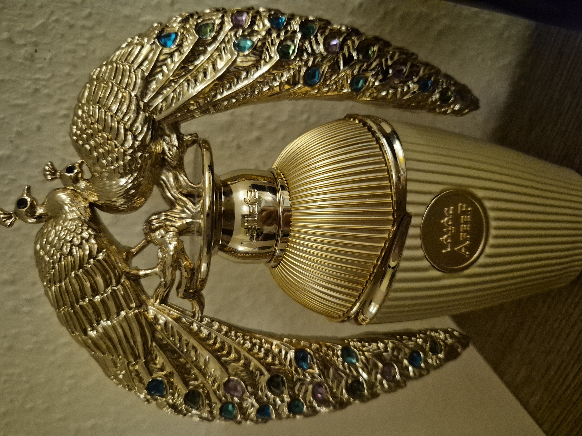 This is a close-up, eye-level, indoor, medium shot of a gold perfume bottle. The bottle has a ridged, bell-shaped body with a ridged cap. Two gold peacocks with jewel-toned accents on their feathers are perched on top of the bottle, facing each other. The peacocks' eyes are small black beads. The bottle has an oval-shaped gold emblem near the bottom with Arabic script. The background is a textured, neutral-colored wall. The lighting is warm and casts shadows, highlighting the metallic sheen of the bottle and peacocks. The image is sharply focused on the perfume bottle. There are no people or animals visible in the scene, and no recognizable landmarks or environmental features that would suggest a specific city or location.