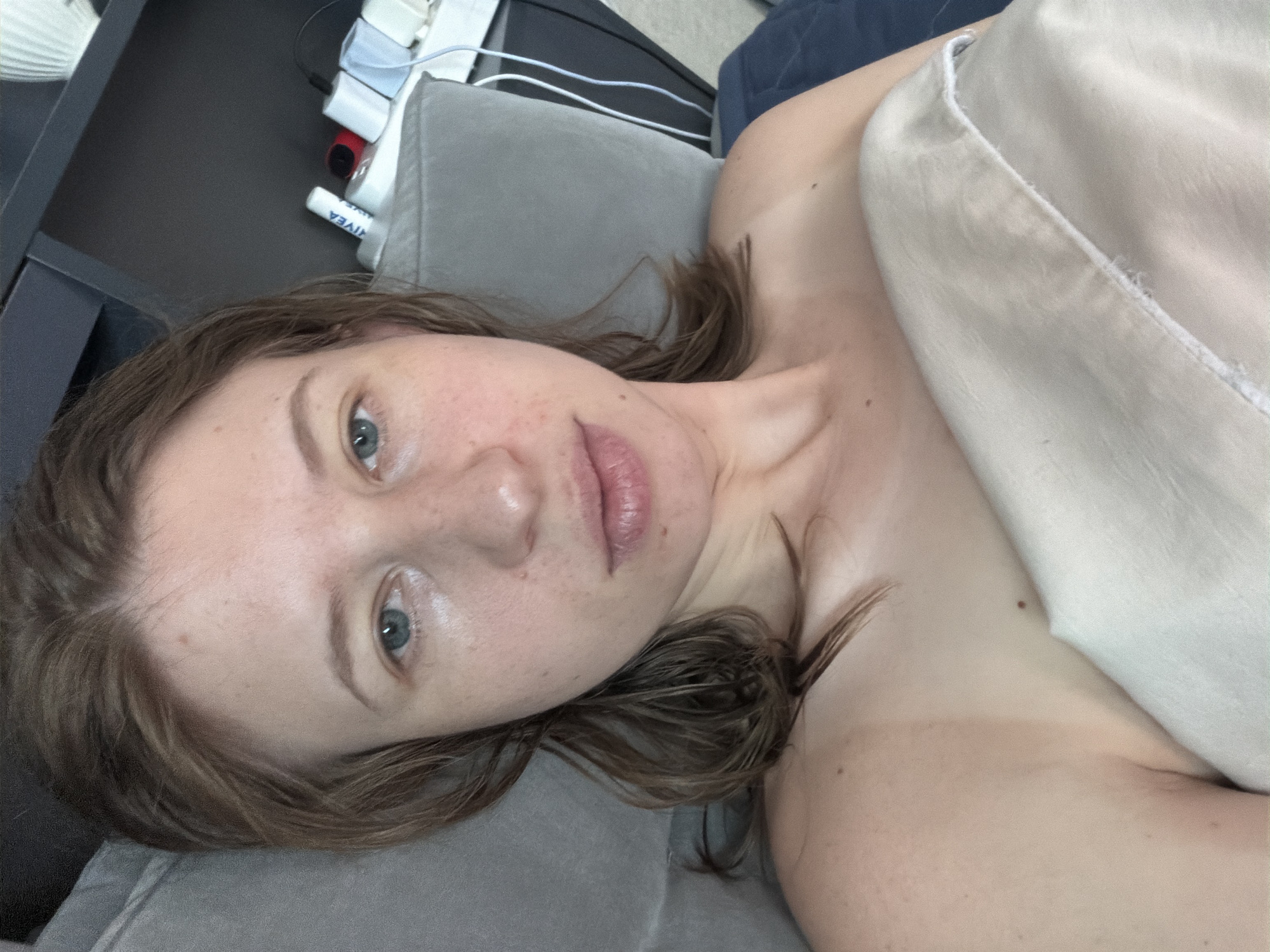 The image shows a close-up, selfie-style view of a fair-skinned woman with light brown hair, lying down and covered by a light-colored blanket. She is looking towards the camera with a neutral expression. Her left eye is visible, along with part of her nose and mouth. The lighting is soft, and the background appears to be indoors, possibly a bedroom or living area, with a dark shelf unit visible to the right, holding several electronic charging devices. There is a hint of a tan line on her shoulder. The image does not contain any recognizable landmarks or indicators to suggest a specific location within Dongducheon-si, South Korea.