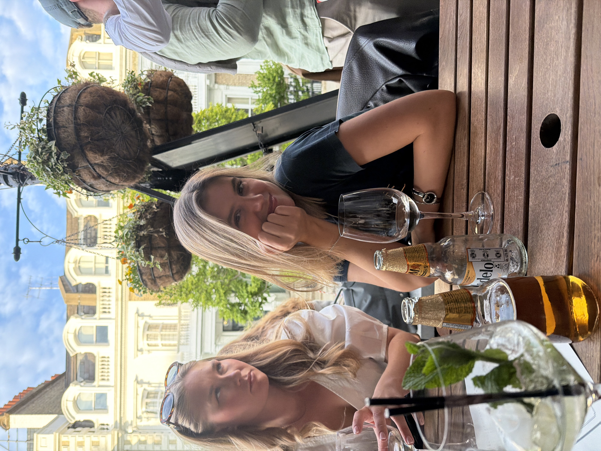 This media file captures an outdoor social gathering, likely at a pub or restaurant patio in London, characterized by its classical architecture and clear sky. The scene is bright, suggesting daytime, possibly late afternoon.
In the foreground, a slatted wooden table is central, holding various drinks. Two women are seated, engaged in the activity. The woman on the left, with long blonde hair, is looking directly at the camera with a friendly smile, her chin resting on her hand. She is wearing a dark t-shirt and has a silver watch on her wrist. Next to her, a clear wine glass stands empty.
To her right, another woman with long blonde hair is partially visible, looking down or away from the camera. She wears a white top, and sunglasses are perched on her head. Her hands are near a tall glass containing a clear drink with ice and mint leaves, resembling a mojito.
On the table between them are several bottles. One is a clear glass bottle with a gold-foiled neck, clearly displaying the brand "Modelo" and the Spanish word "ADVERTENCIA" (warning) on its label. Another bottle, amber-colored and also with a gold-foiled neck, has the text "VALOREM" visible on its label.
The background features grand, light-colored buildings with arched windows and ornate detailing, typical of London's architectural style. Overhead, hanging baskets overflowing with green foliage and small yellow flowers are suspended, adding to the pleasant outdoor ambiance. A clear blue sky with a few wispy clouds is visible above the buildings. A man wearing a cap and a light green shirt is partially visible in the upper background, seated at another table.
The overall atmosphere appears relaxed and convivial.