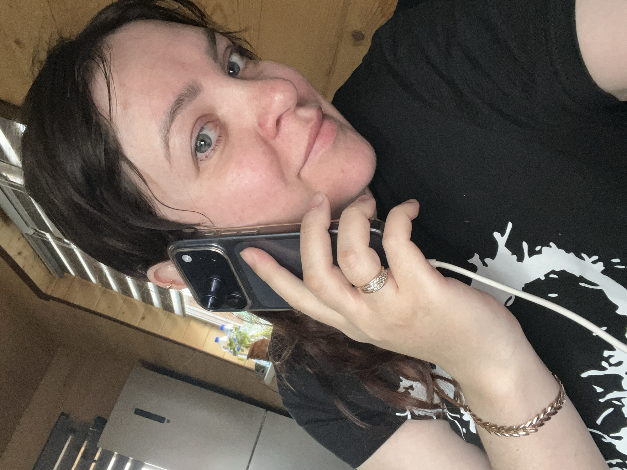 A medium-shot, angled, indoor portrait captures a woman holding a black smartphone to her left ear. She appears to be on a call, and a white charging cable is connected to the phone, visible near her wrist. She is wearing a black t-shirt with a white graphic print. A gold-colored bracelet adorns her left wrist, and a multi-stone ring is on her ring finger. The woman has light skin, dark hair, and blue-grey eyes. The background includes what appears to be a wooden wall, a window, and part of a refrigerator. Specific location details regarding Protasy, Russia are not discernible from the image.