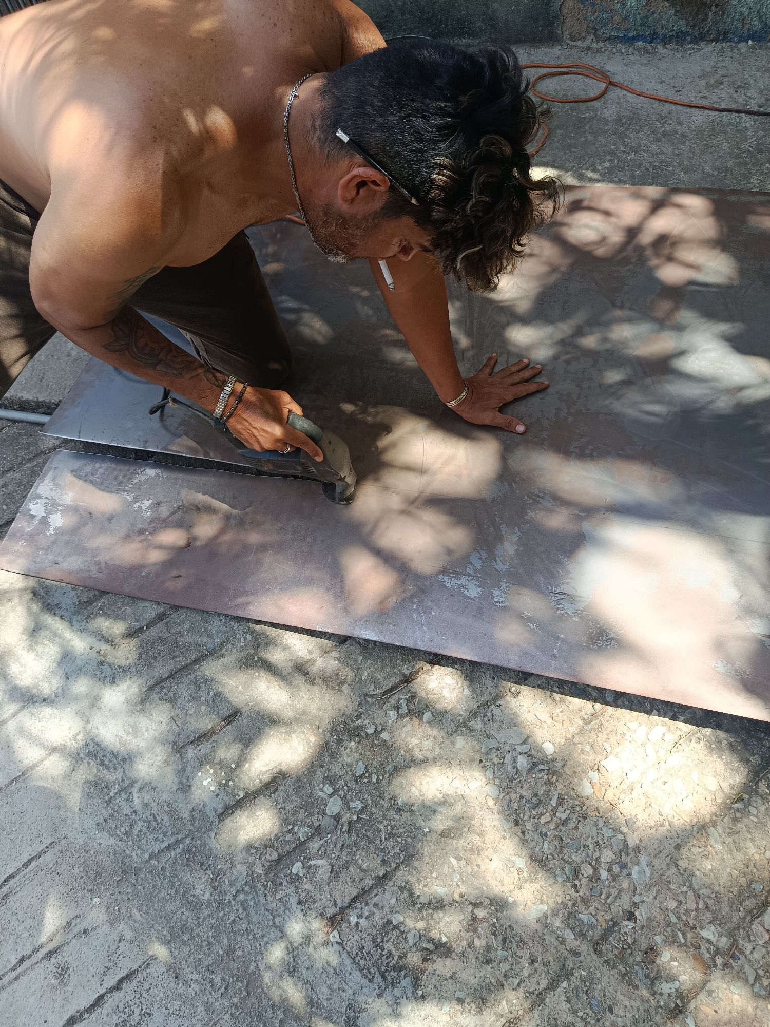 A shirtless man with dark hair and a cigarette in his mouth is kneeling on a cracked concrete surface. He is using a power tool with a blue handle on a large sheet of metal. He is wearing bracelets on his left wrist and has a tattoo on his left arm. The scene is outdoors in bright sunlight. The location is Juan Griego, Venezuela.