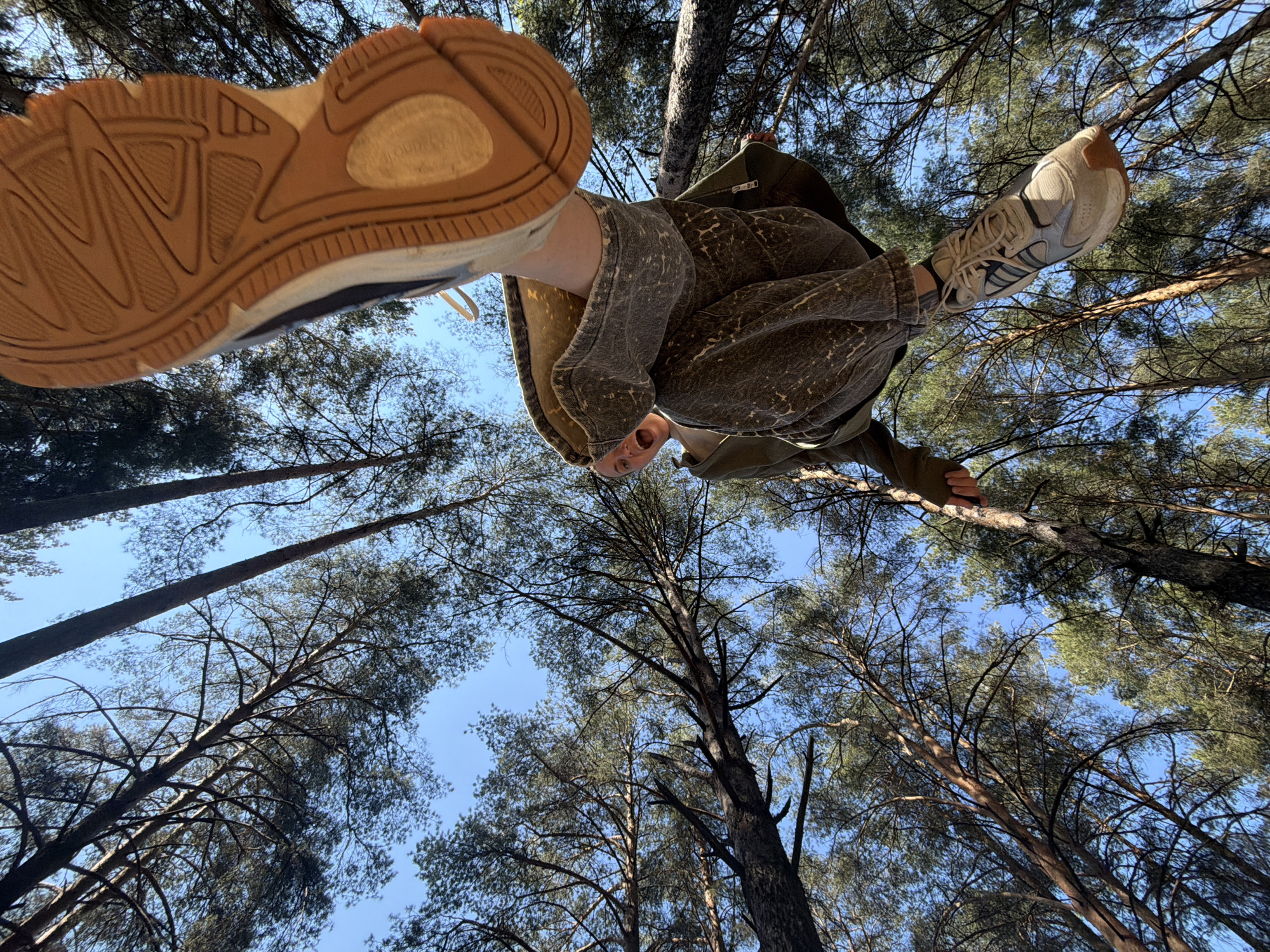 A low-angle shot shows a person from the feet up against a backdrop of tall pine trees. The person is wearing a distressed green outfit. The person's mouth is open and appears to be shouting or in surprise. Their feet are pointing towards the camera. The surrounding environment is a dense forest of pine trees, stretching upwards towards a blue sky visible through the branches. The trees suggest the location is in a natural area, likely a park or forest on the outskirts of Chaikovsky, Russia. The shoes appear to be sneakers with a heavy tread.