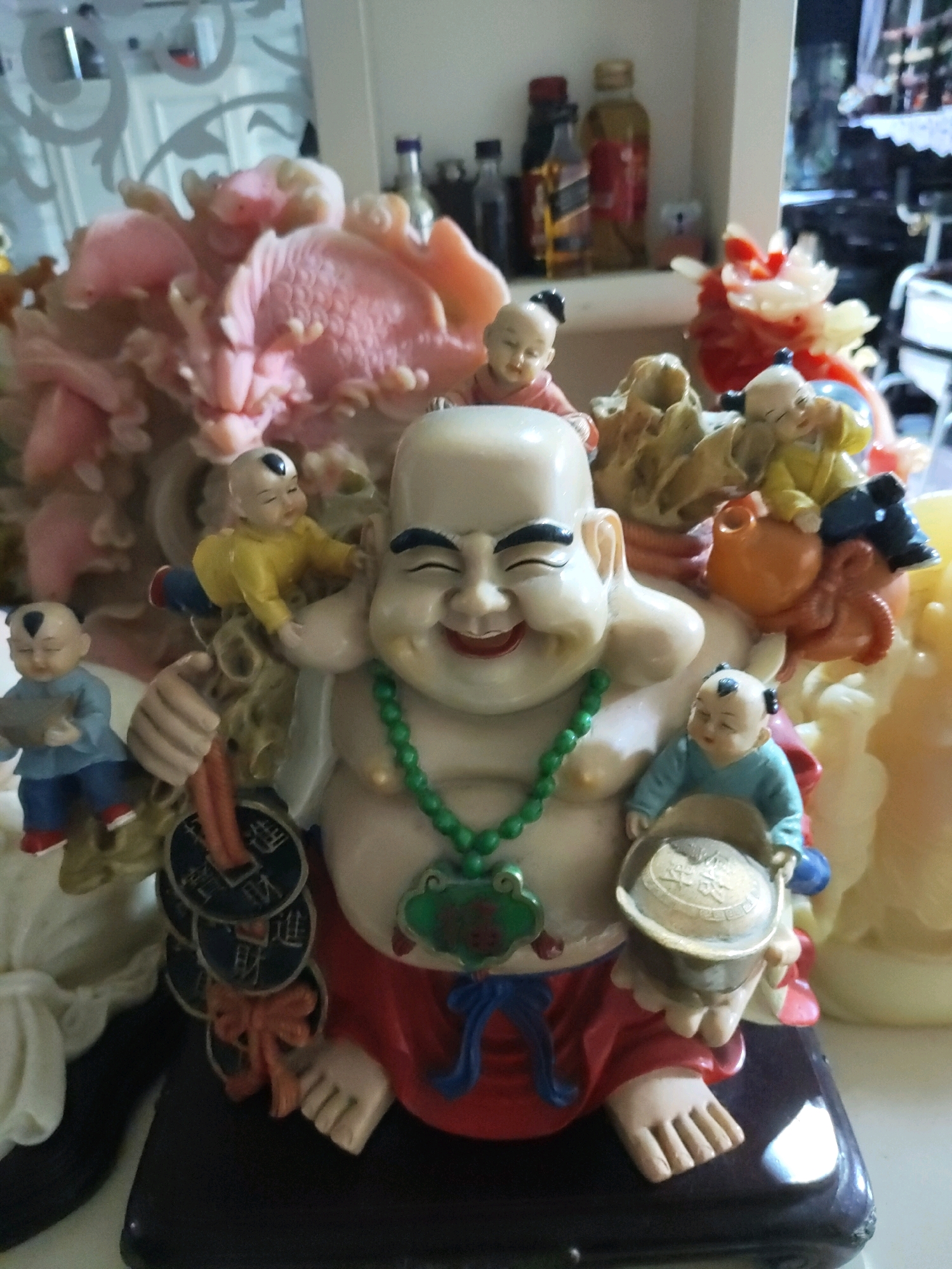 The image captures a close-up view of a decorative figurine, likely a Budai (Laughing Buddha) statue, adorned with several smaller figures. The main figure, a large, smiling Budai, is seated and depicted with rosy cheeks, closed eyes, and a jovial expression. He wears a green beaded necklace and a red garment. Surrounding him are five child-like figures, some of whom are interacting with him. One child is perched on his shoulder, another is positioned on his lap holding a shallow bowl, and two more are at his feet. One of these children, dressed in a blue top and trousers with red shoes, holds a grey object. Another child, in a yellow tunic, is leaning towards the Budai. The figurine is placed on a dark, glossy surface that appears to be a shelf or stand. In the background, out of focus, are shelves with various items, including bottles and other decorative objects, suggesting an indoor setting. The overall scene is brightly lit, highlighting the colorful details of the figurines. The location is described as New Taipei City, Taiwan.