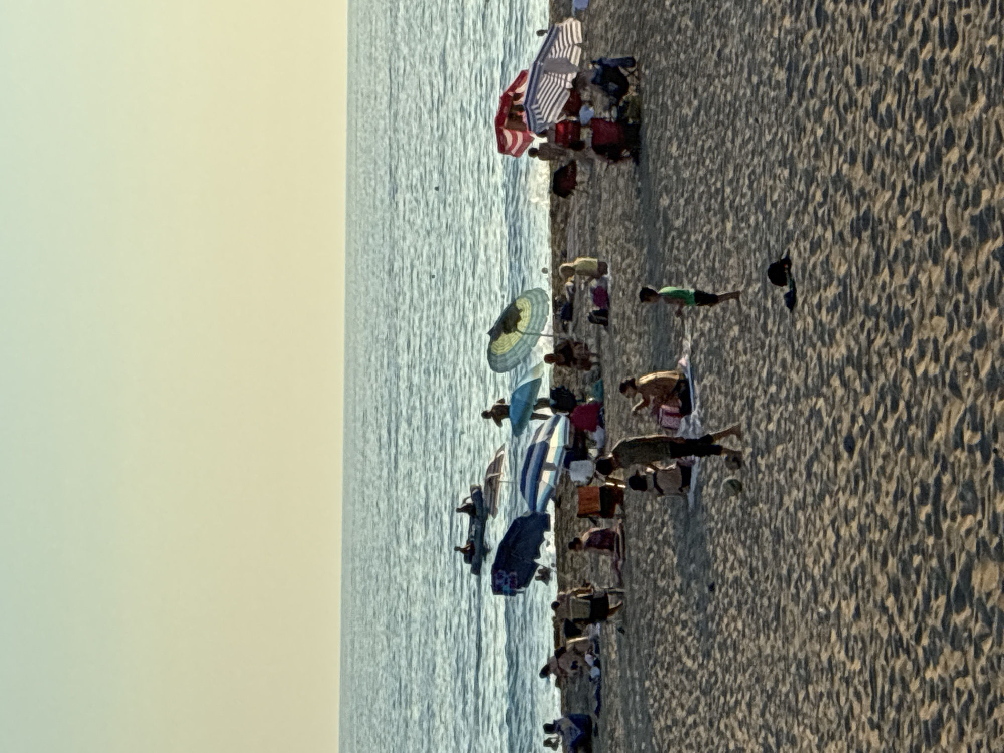 The image depicts a crowded beach scene with people enjoying leisure activities on the sand and in the shallow water. The ocean is calm with gentle waves. Several beach umbrellas, some striped and others solid colors, provide shade for beachgoers. People are seen sitting, lying down, and walking on the beach. In the water, a small boat with a few occupants is visible. The sand is textured with visible footprints and shadows from the umbrellas. The sky above is a pale, gradient hue, suggesting either early morning or late afternoon. The overall atmosphere is relaxed and summery. The location is identified as Alanya, Turkey.