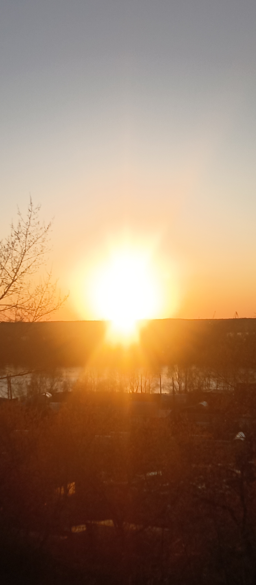 The image depicts a bright sunset. The sun is positioned near the horizon, emitting a strong, radiant light. The sky transitions from a pale gray to an orange hue as it approaches the horizon. Silhouetted trees and shrubs are visible in the foreground. In the middle ground, there is what seems to be a body of water. On the horizon are industrial cranes. The location appears to be on the outskirts of a city.