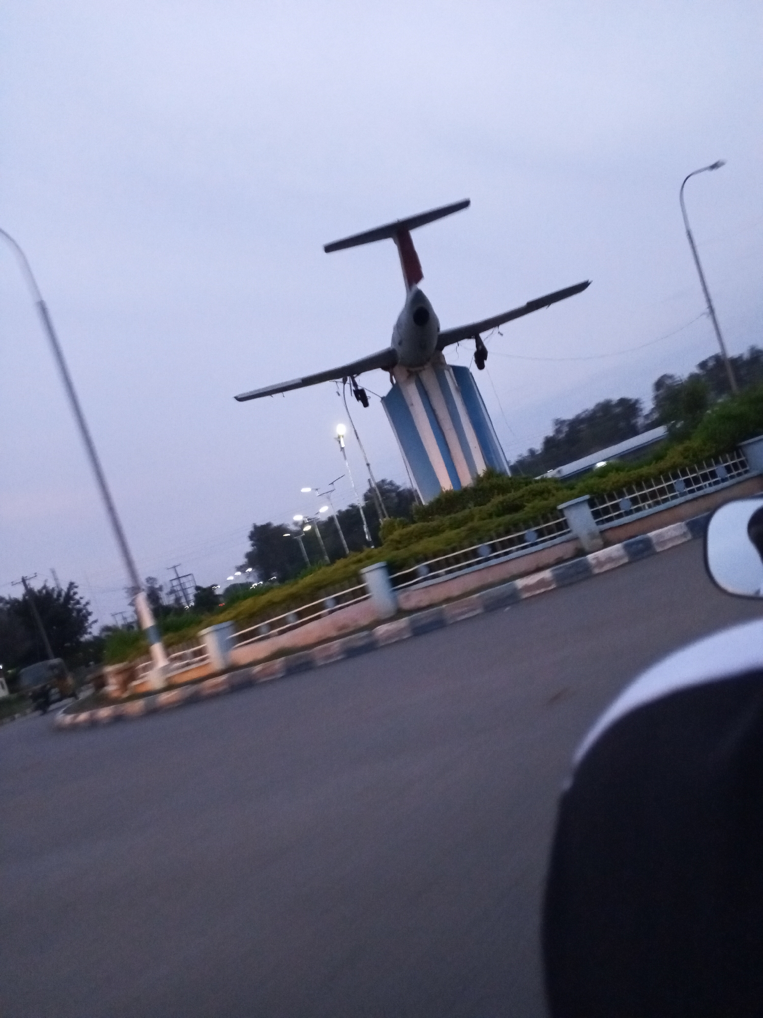 An airplane monument stands prominently in the city of Jos, Nigeria. The monument is elevated on a striped blue and white cylindrical base. The airplane itself is gray with a red stripe on its tail and appears to be a model jet. Surrounding the monument is a low green hedge bordered by a fence with white and black patterned posts. A paved road curves around the monument, with a visible portion of a motorcycle's side mirror in the foreground, indicating the image was taken from a moving motorcycle. Streetlights are visible in the background, and the sky is overcast.