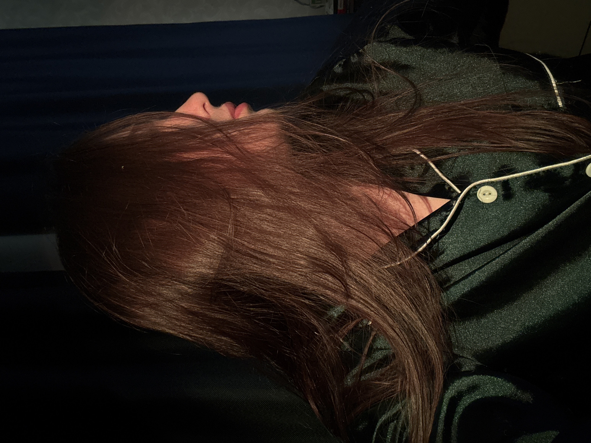 A person with long, auburn hair lies on their side on a dark blue bed. They are wearing a dark green satin pajama top with white piping and white buttons. The scene is dimly lit. The location is unknown, as there is no information to indicate the location is Chaikovsky, Russia.