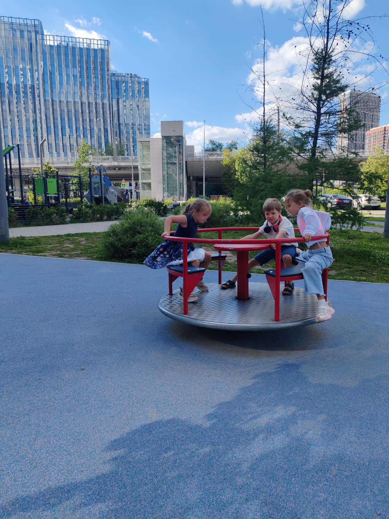 In Moscow, Russia, three children are playing on a red merry-go-round in a playground with a blue rubberized surface. Two girls and one boy are on the merry-go-round. One girl on the left wears a dark blue dress with a floral pattern and white sneakers. The boy in the center is wearing a white t-shirt with red headphones around his neck, blue shorts, and dark sandals. The girl on the right is wearing a white crop top, light blue jeans, and white sneakers. In the background, there are modern glass buildings, trees, and a playground structure. The sky is blue with scattered white clouds.