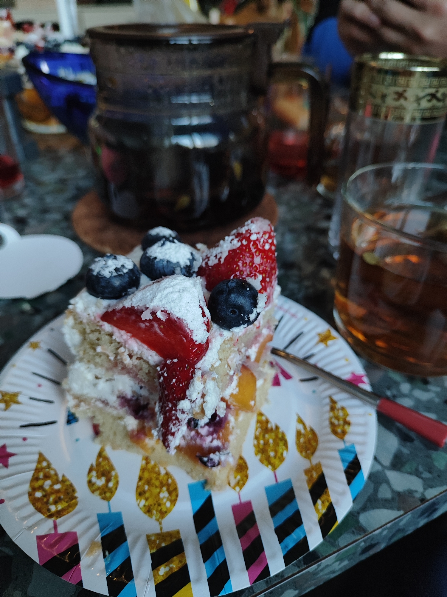 A close-up, eye-level shot captures a festive scene centered around a slice of cake. The main subject is a generous slice of layered cake served on a white paper plate, adorned with colorful birthday candle illustrations and small stars. The cake itself features light-colored sponge layers, white whipped cream or frosting, and is topped with fresh strawberries, blueberries, and a dusting of powdered sugar. Peaches or another yellow fruit are visible within the cake layers, adding to its fruit content.
In the background, several dark-colored glass items are visible, including a large pitcher or teapot and a smaller glass containing a dark amber liquid, possibly tea, with what appears to be cardamom pods floating in it. Another glass with an ornate golden rim is partially visible. A small spoon or knife with a red handle rests on the plate next to the cake. A hand is briefly seen in the upper right background, indicating the presence of people, though no faces are visible. The setting appears to be indoors, possibly on a patterned countertop or table. The use of a birthday-themed plate strongly suggests a celebration, likely a birthday party or a similar festive gathering. No discernible text is visible in the image.