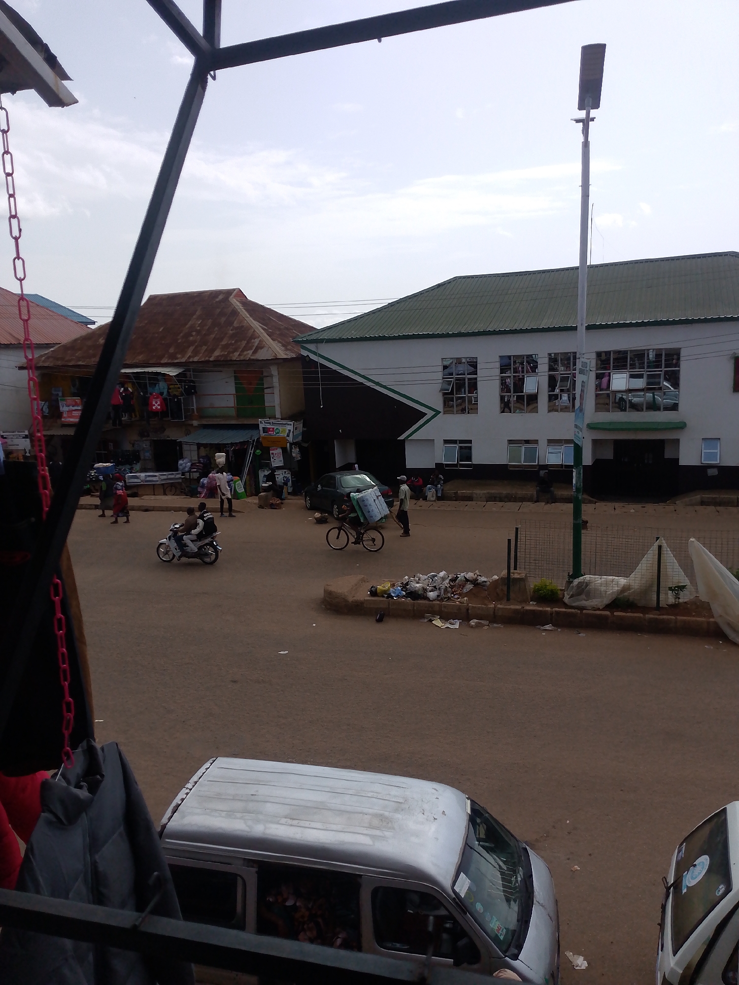 The image captures a street scene in Bukuru, Nigeria. A street light stands tall on the right side of the frame. In the foreground, a silver van is parked. Further down the street, another white van is visible. People are walking and riding on a motorbike, with one person pushing a bicycle laden with goods. A market with stalls selling various items is set up along the road. Buildings with corrugated iron roofs and white exteriors line the street. The road is paved with dirt and littered with trash. The sky is overcast.