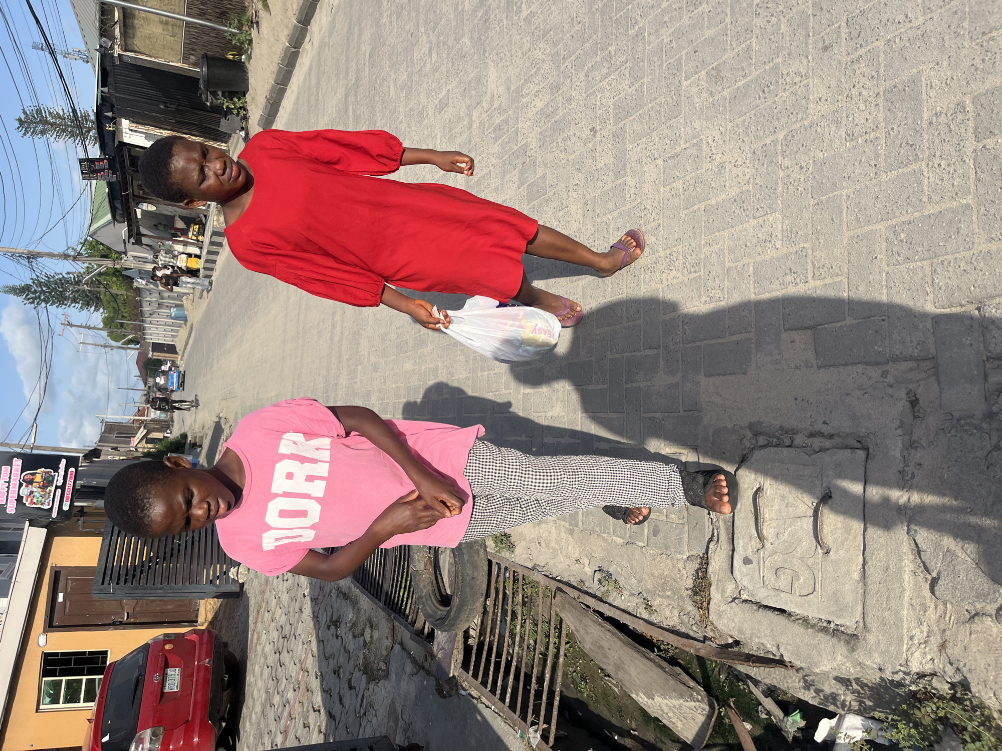Two young women stand in Iranla, Nigeria. One, in a red dress, walks away on a brick road, carrying a white plastic bag. The other, in a pink shirt and checkered pants, stands near a rusty grate and tire on a littered street. A building and red car are visible in the background.